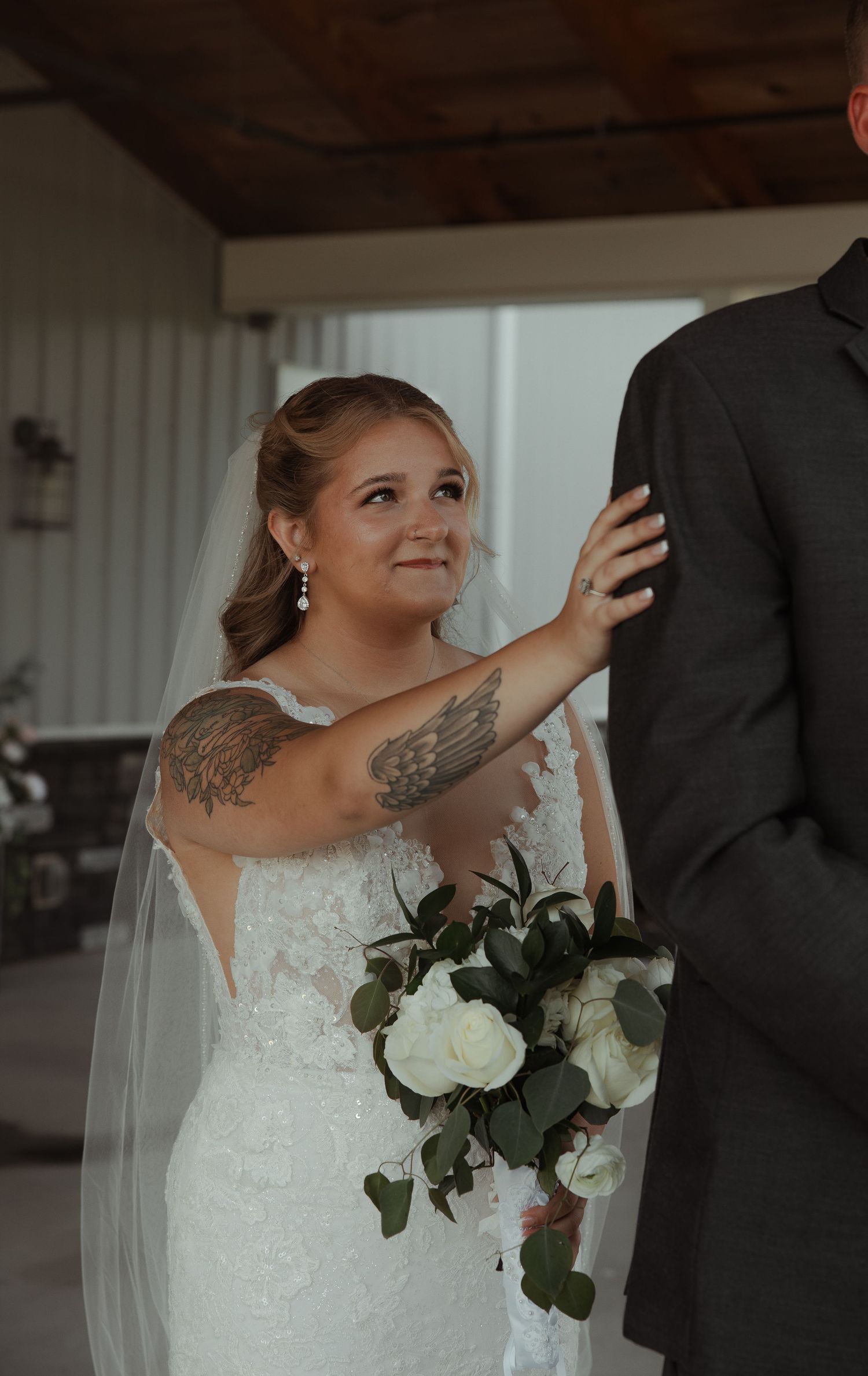 Emotional bride in lace wedding dress with angel wing tattoos holding white bouquet during ceremony.