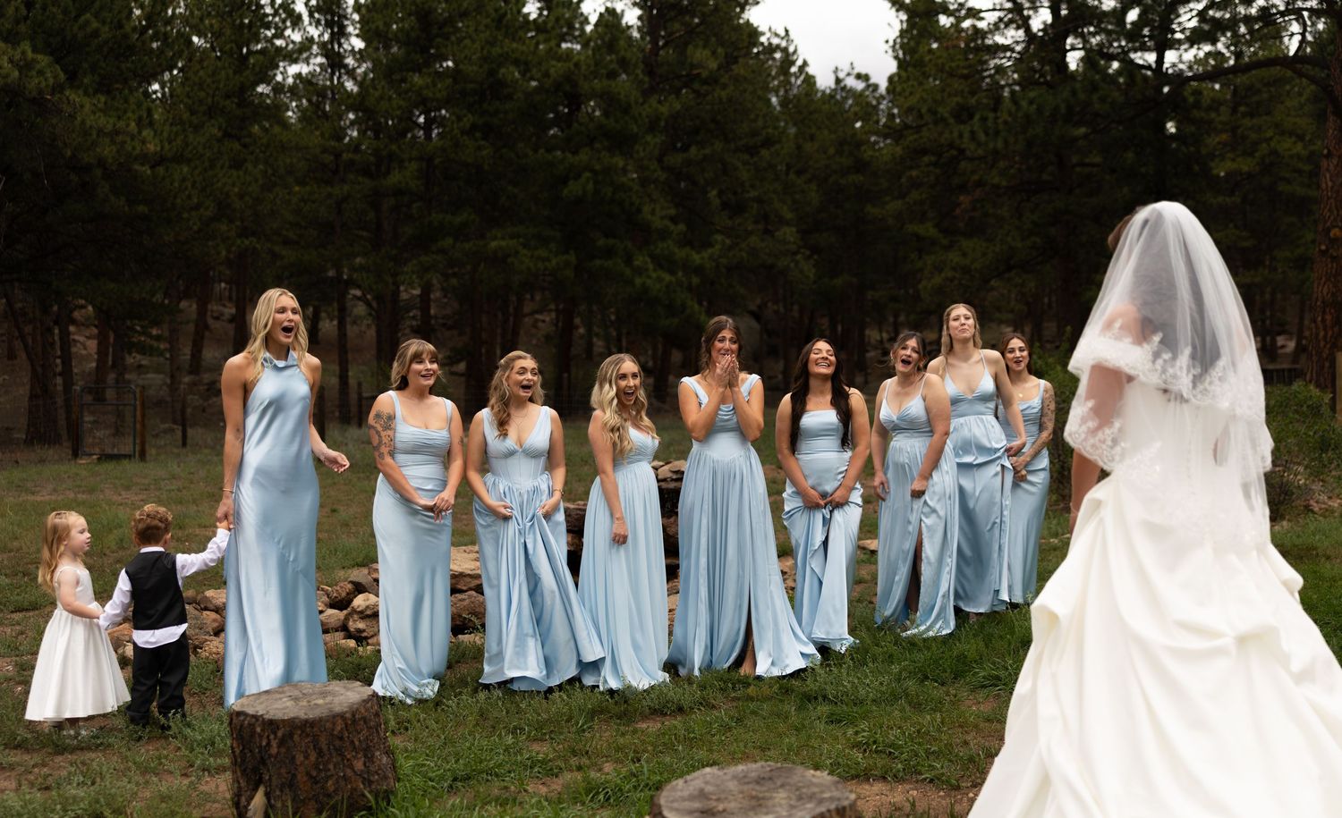 Bride in white gown and veil stands before bridesmaids in light blue dresses at an outdoor forest wedding ceremony.