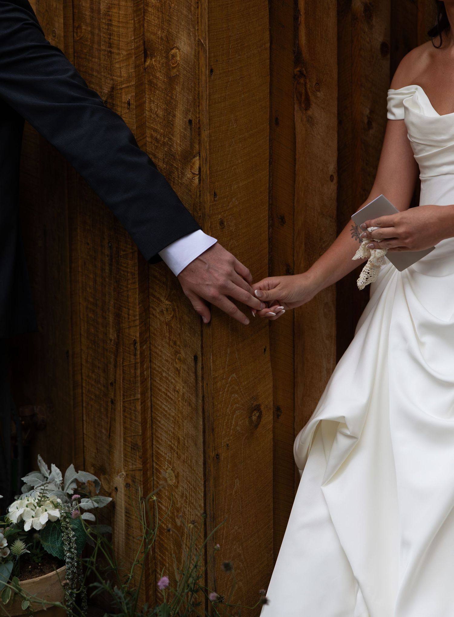 Bride and groom holding hands in front of rustic wood wall, bride in white off-shoulder gown holding vows card.
