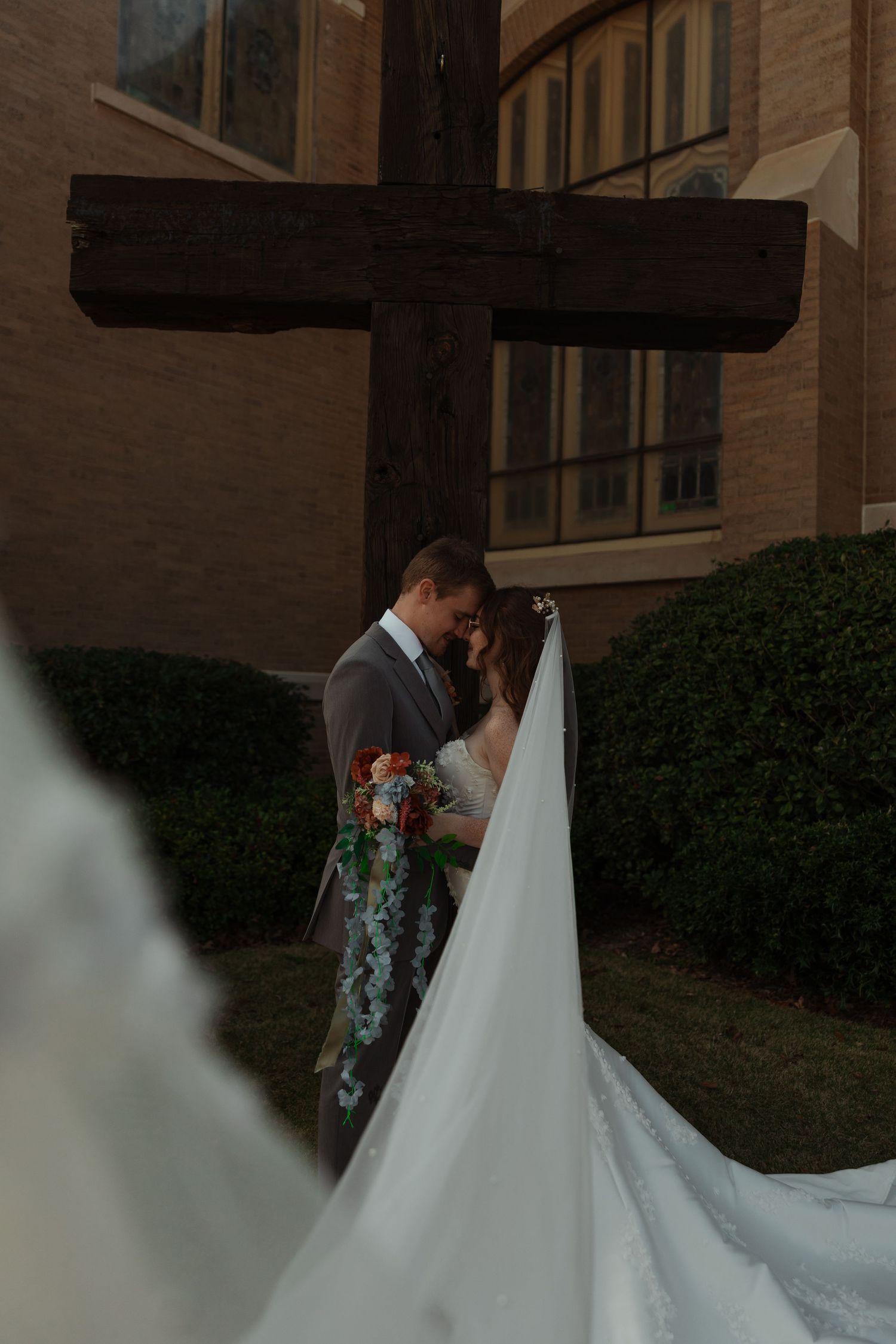 Bride and groom share a romantic kiss beneath a large wooden cross outside a brick church.