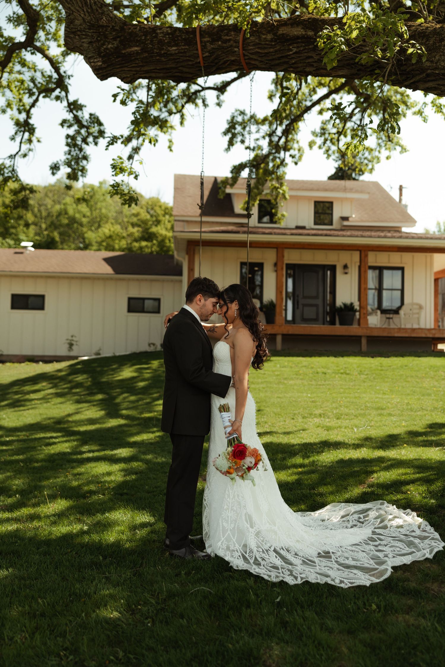 Bride and groom sharing intimate moment under oak tree with farmhouse venue in background.