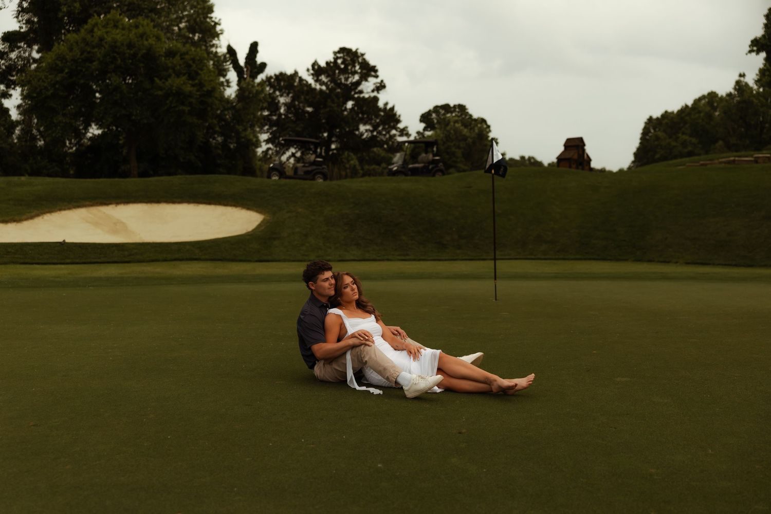 Couple relaxes together on golf course green near flag during casual outdoor engagement photo session.