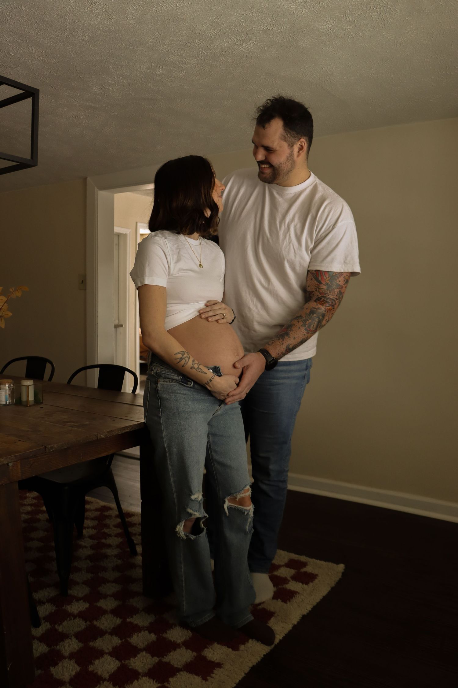 Tattooed couple holding pregnant belly while gazing at each other in warmly lit dining room.