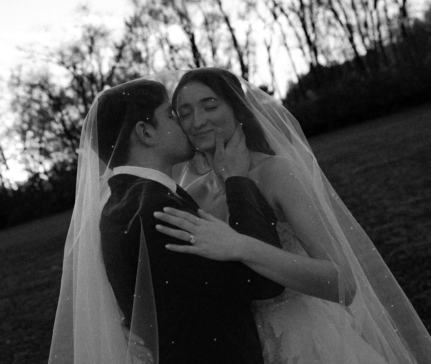 Black and white photo of groom kissing bride's cheek as she smiles under a pearl-dotted veil.