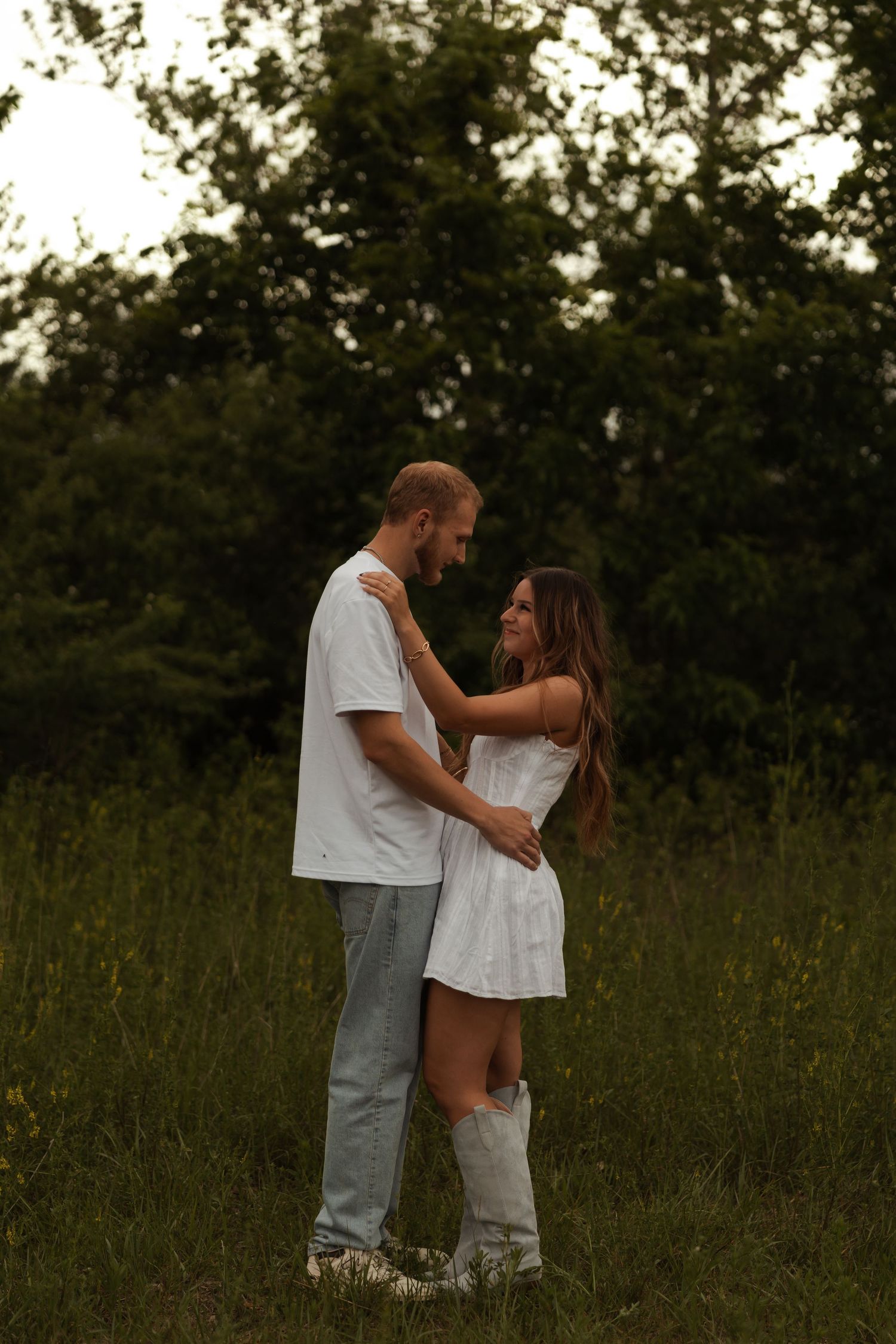 A couple in coordinating white outfits share a tender embrace in a lush green meadow at dusk.