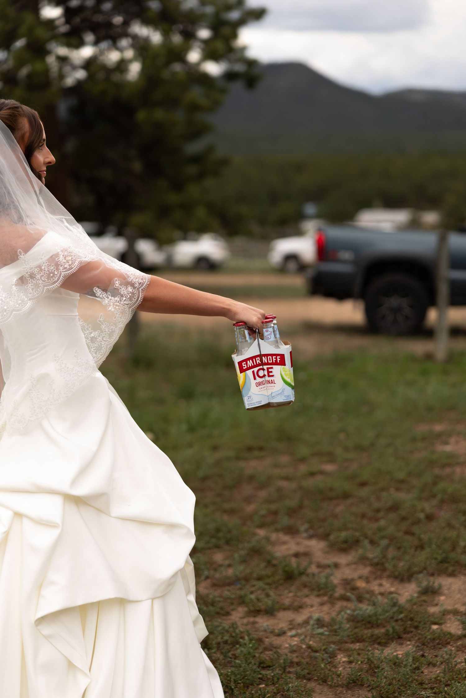 Bride in white wedding gown and veil holding Smirnoff Ice bottles outdoors with mountains in background.