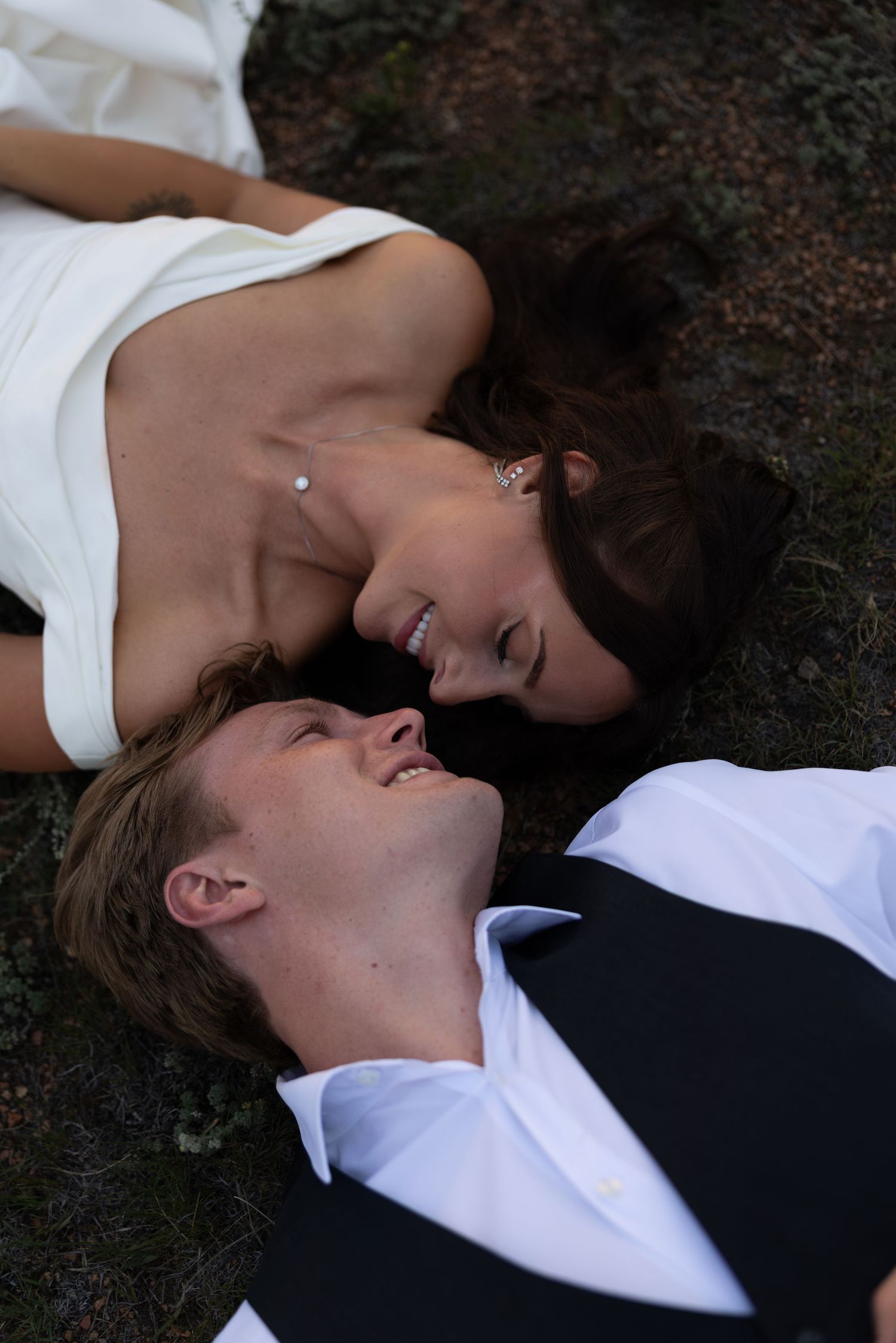 A smiling bride and groom in wedding attire lying on the ground face to face, sharing a joyful romantic moment.