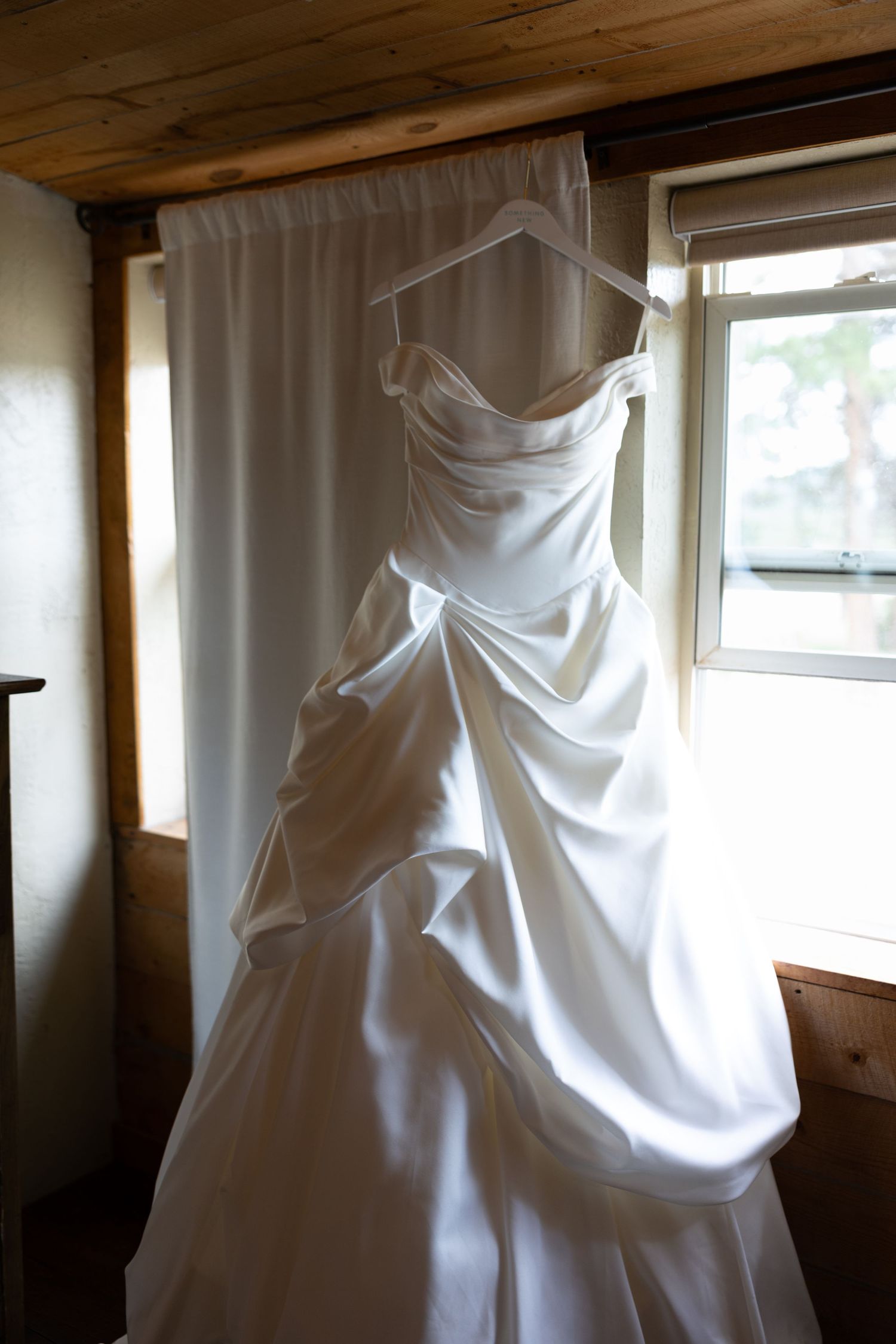 Elegant white satin wedding dress hanging on a hanger in front of a window with soft natural light.
