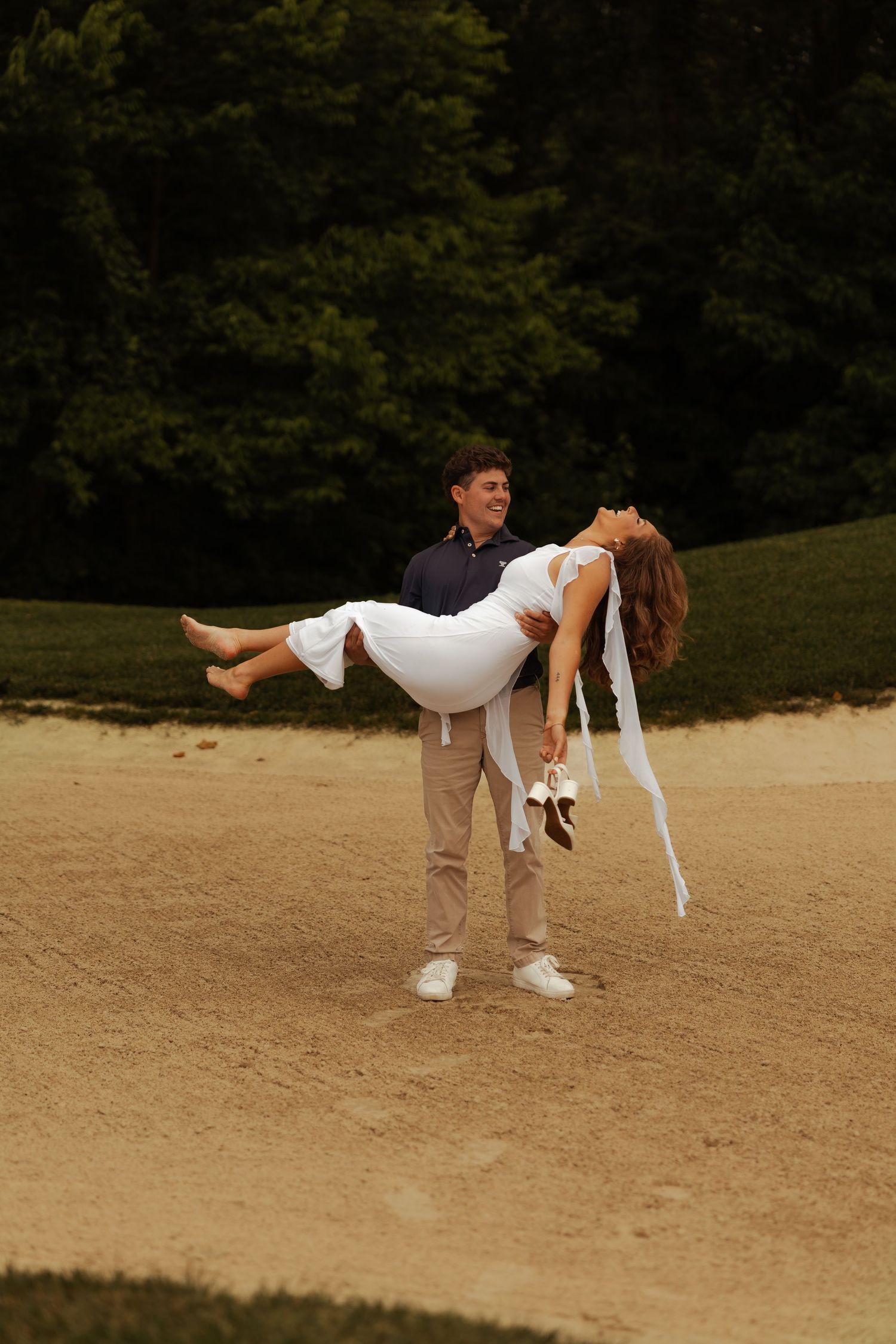 Groom carries barefoot bride in white dress across sandy golf course bunker during engagement photos.