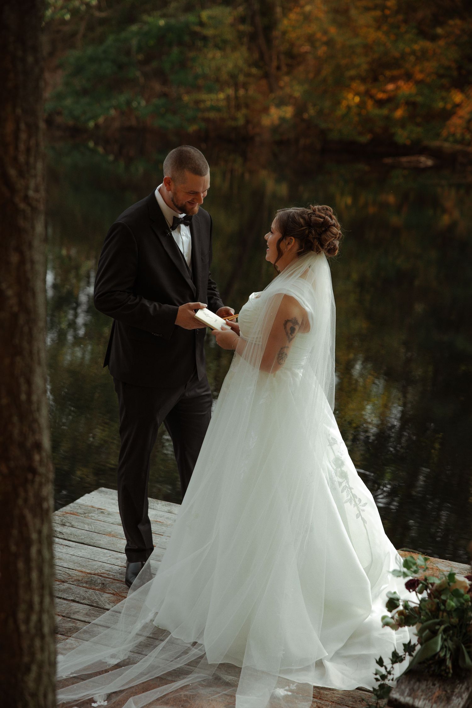 Bride and groom exchange vows on a wooden dock by a reflective lake surrounded by autumn foliage.