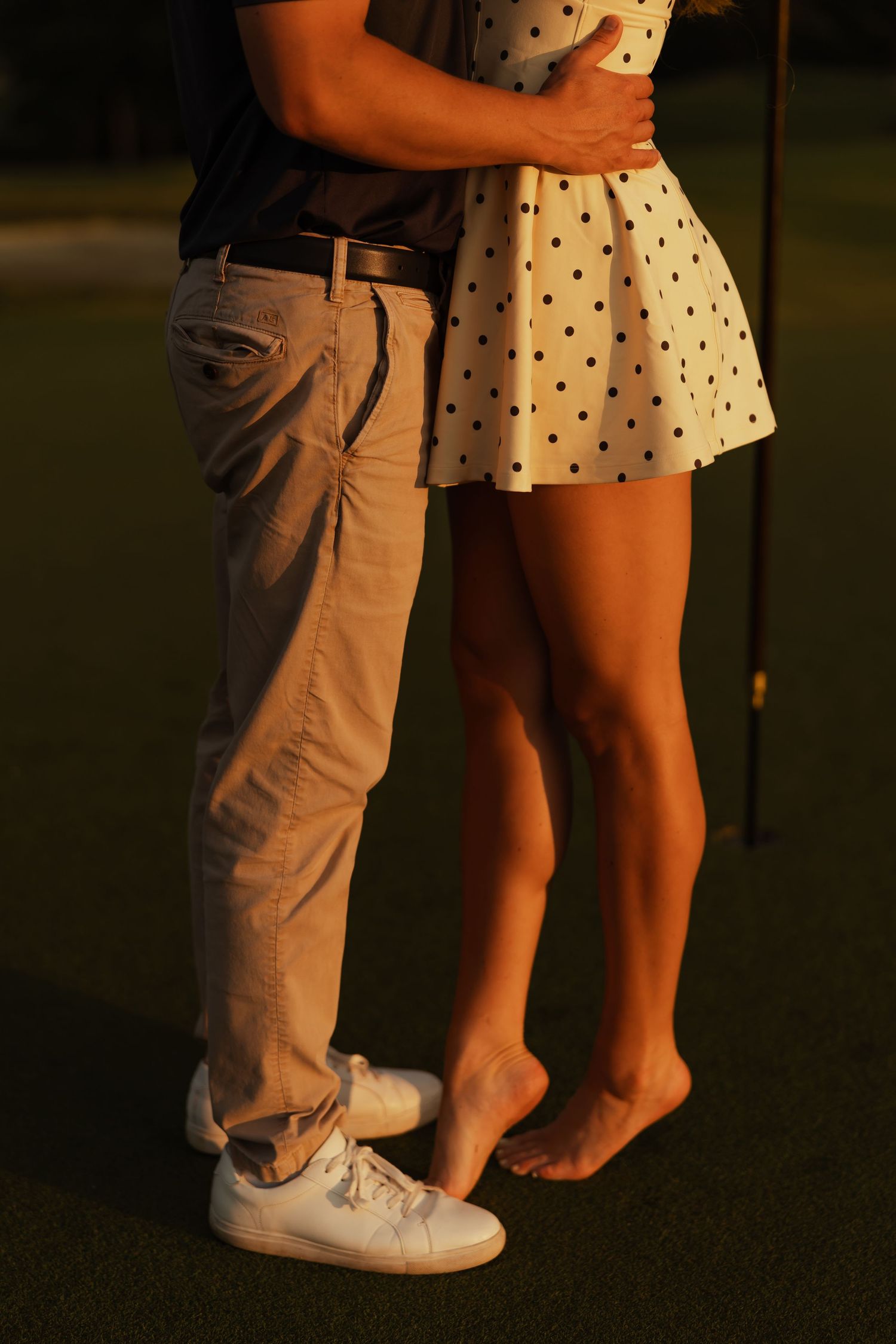 Couple embracing at sunset on golf course, woman in polka dot skirt standing barefoot.
