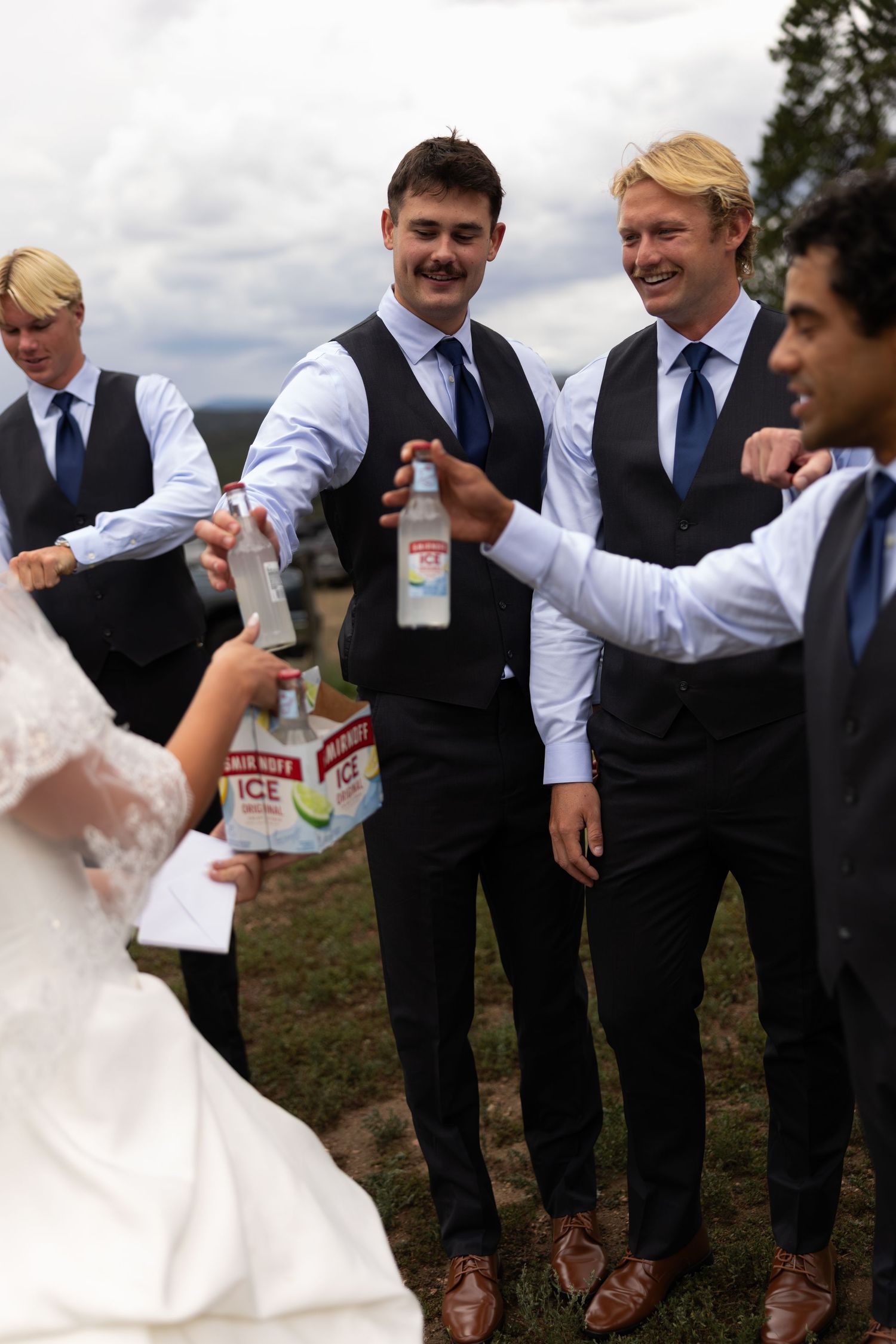 Groomsmen in black vests and blue ties sharing Smirnoff Ice drinks with bride at outdoor wedding reception.