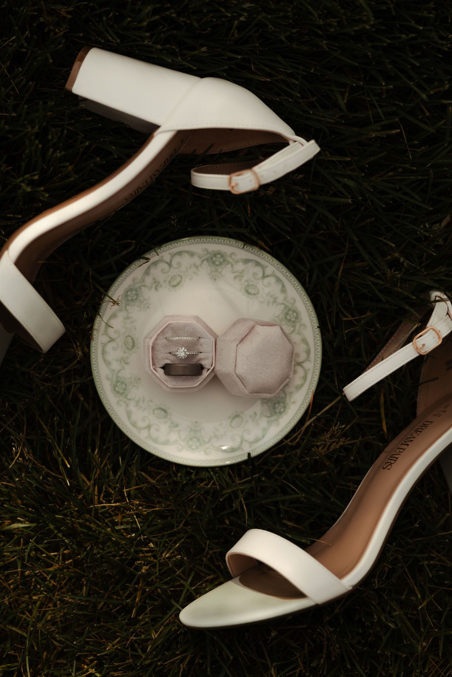 White bridal heels and diamond engagement ring in pink velvet box on decorative plate in grass.