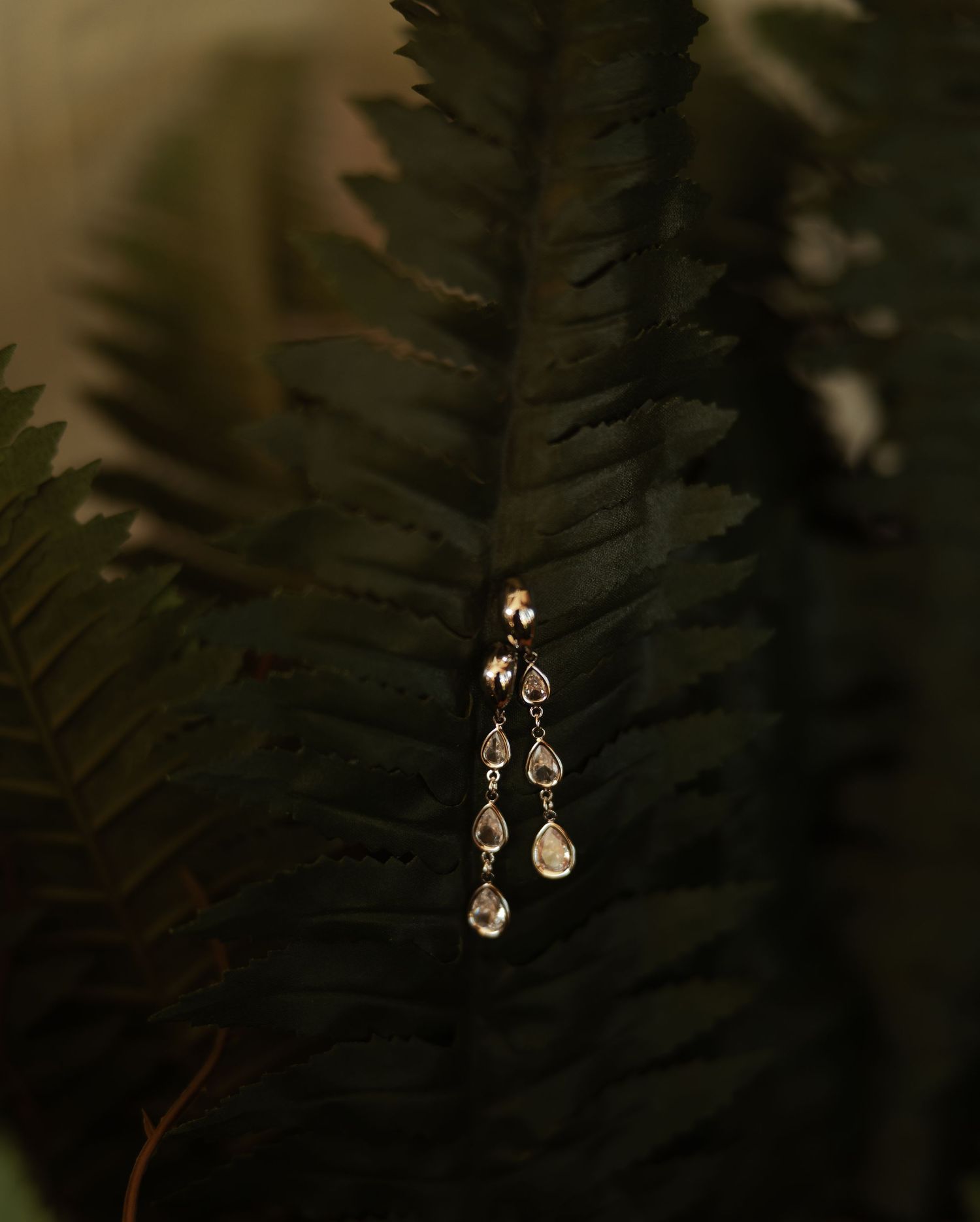 Elegant dangling crystal teardrop earring displayed on dark green fern leaf in moody lighting.