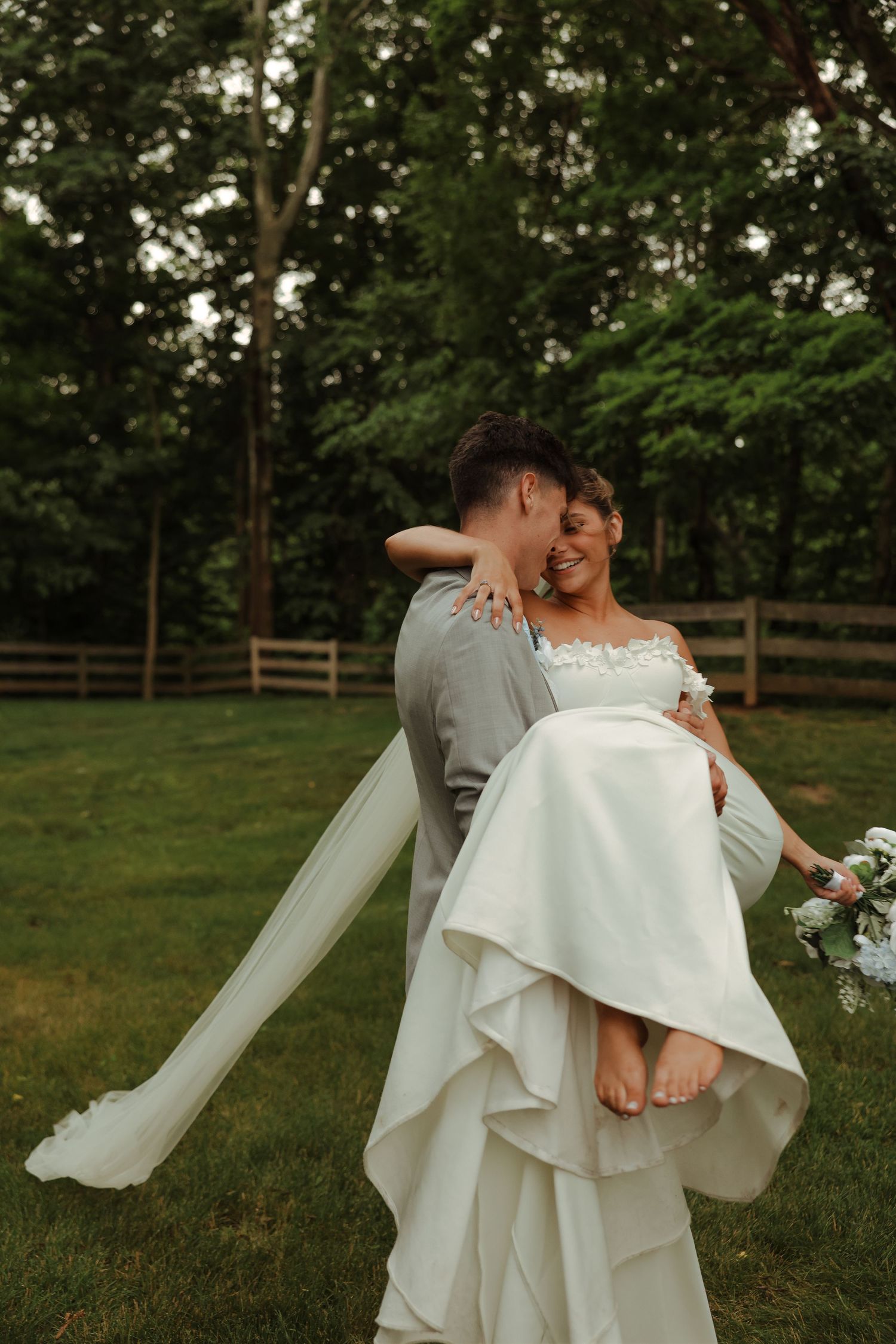 Groom in gray suit lifts laughing barefoot bride in white gown in lush garden setting.
