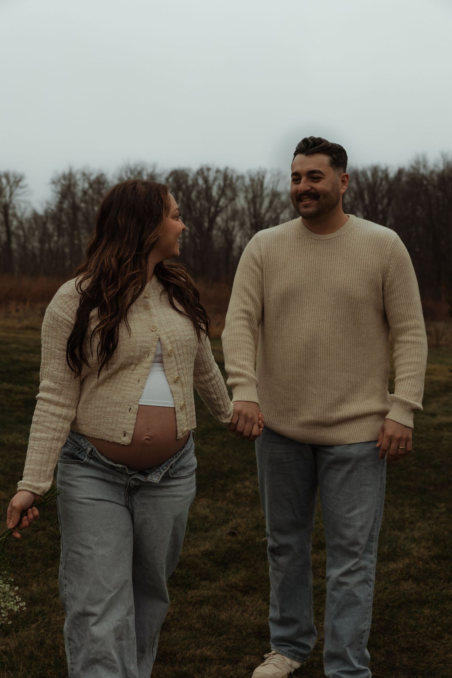 Smiling couple holding hands in open field during casual outdoor maternity photography session.