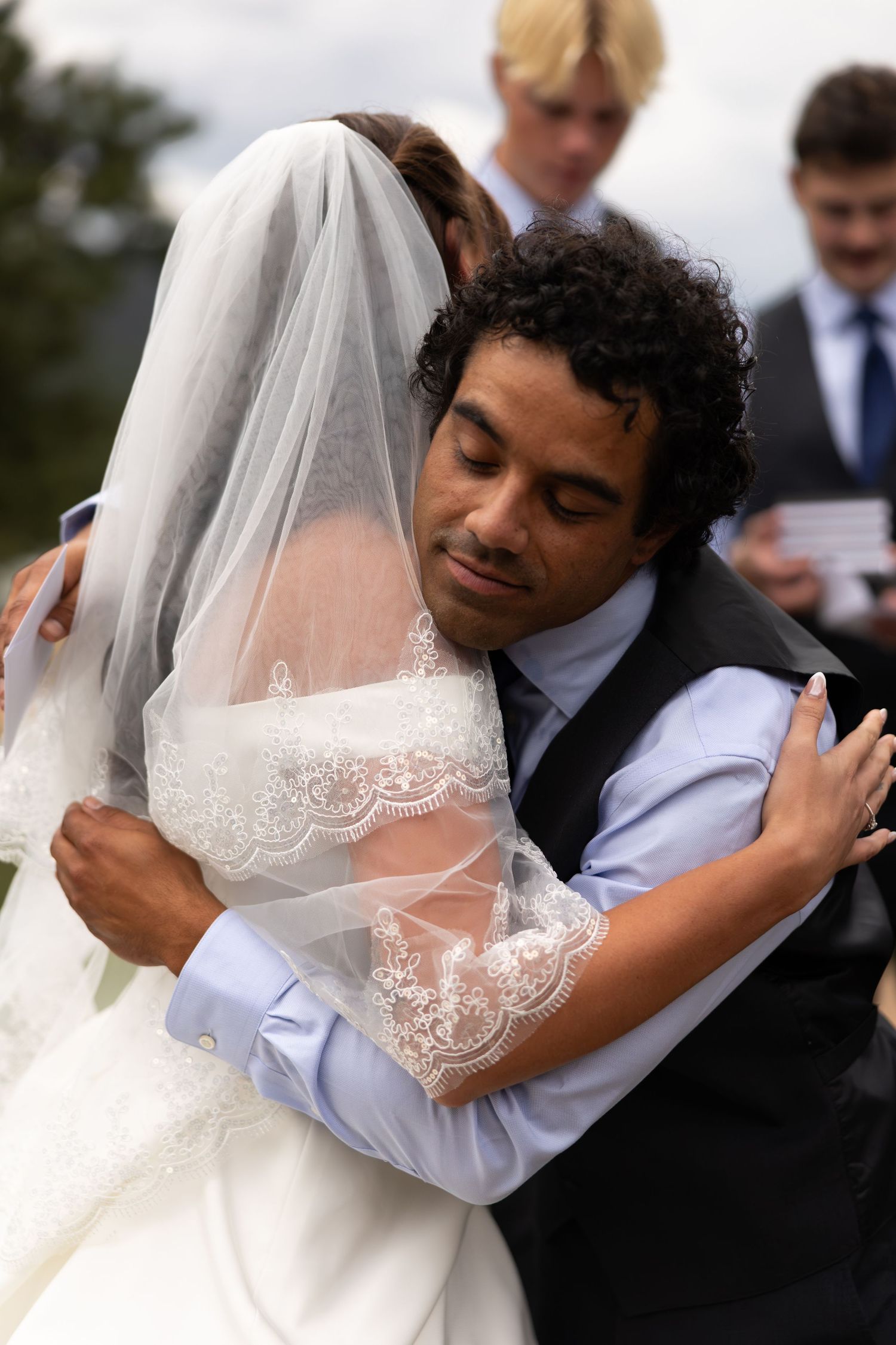 Newlywed couple sharing an emotional hug during outdoor wedding ceremony, bride in lace veil and groom in suit.