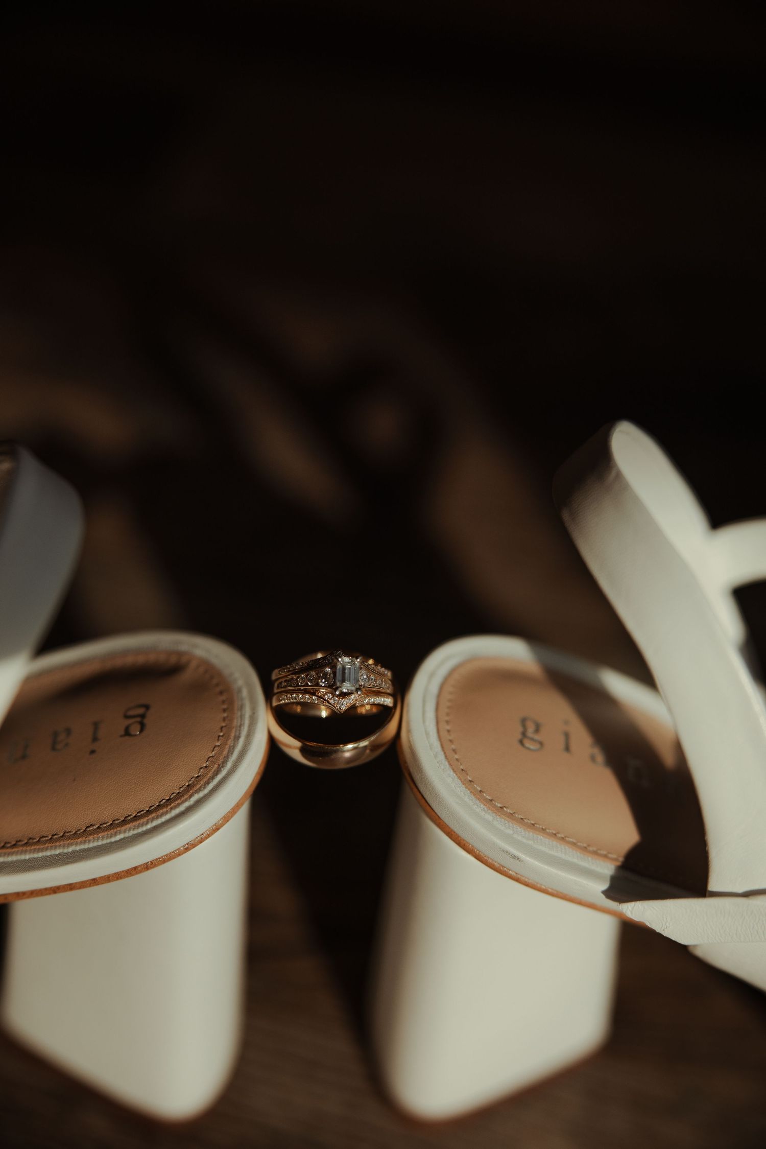 Gold wedding band and diamond engagement ring resting between white bridal heels by Gia.