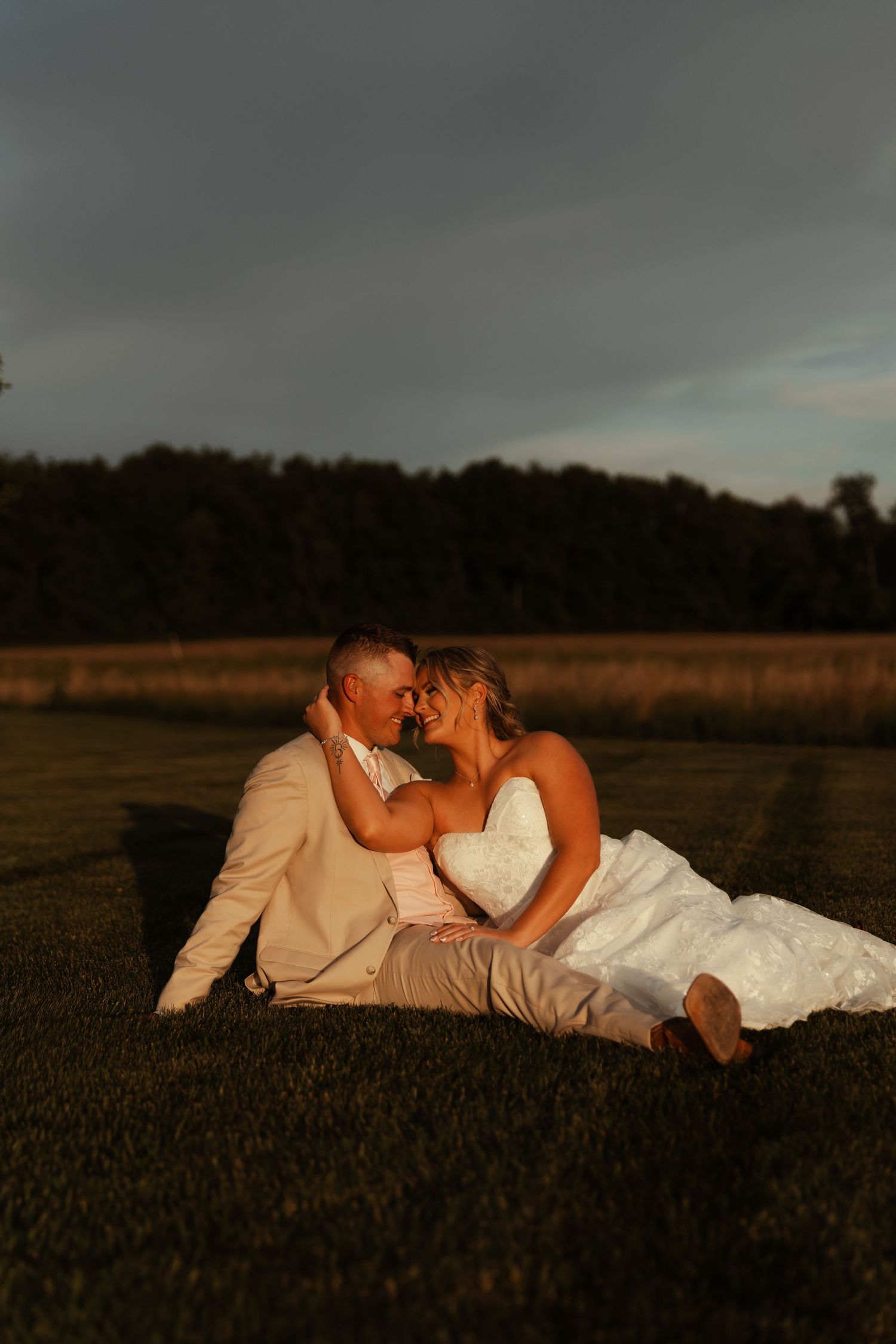 Bride and groom sitting together in a field at golden hour sunset, smiling.