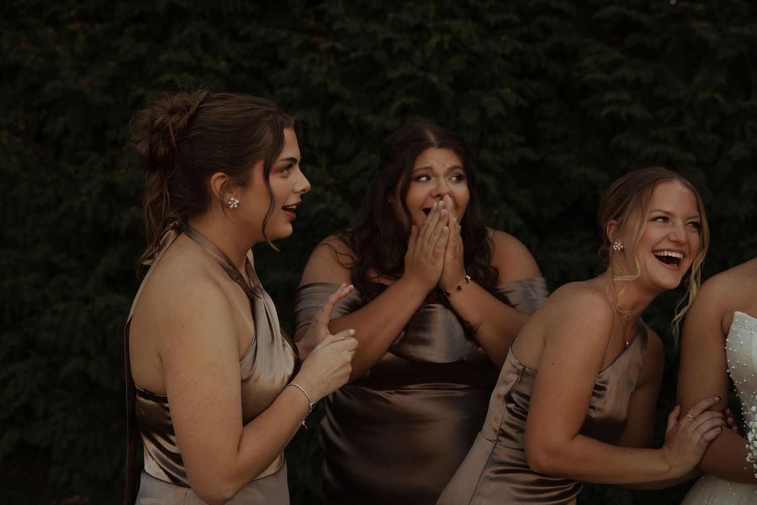 Three bridesmaids in champagne satin dresses reacting with surprise and laughter at a wedding.