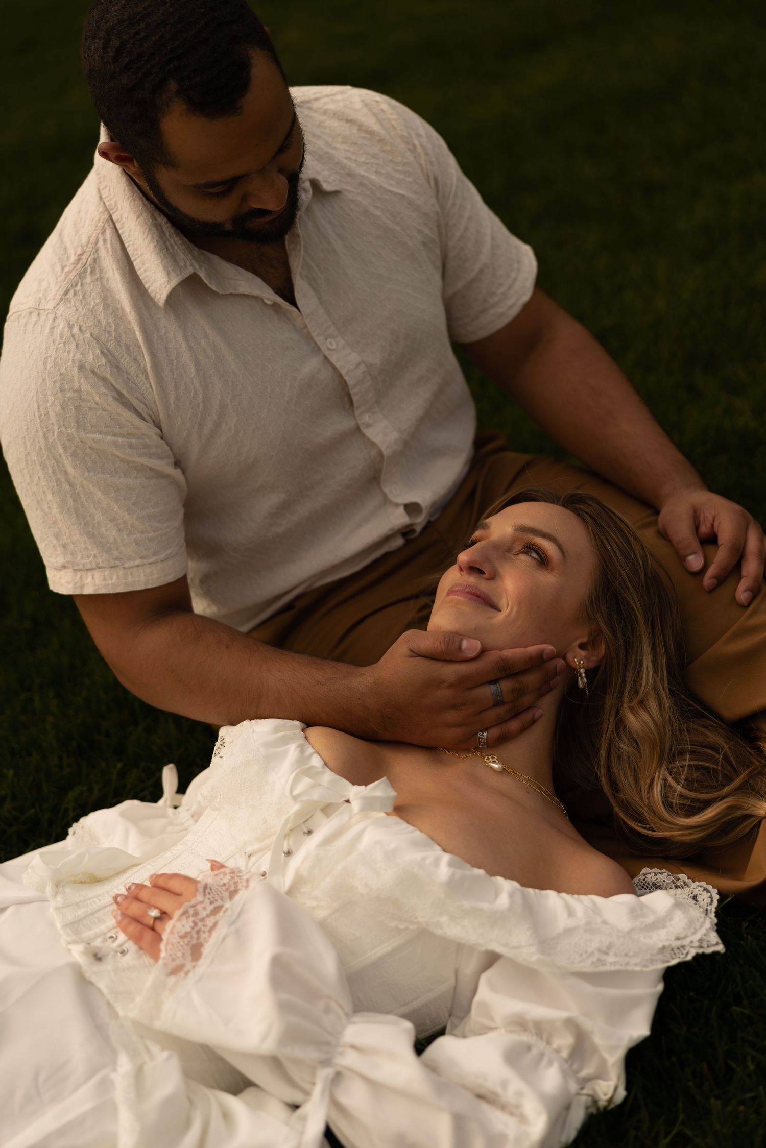 Romantic couple on grass, man tenderly holding womans face as she lies in white wedding dress.