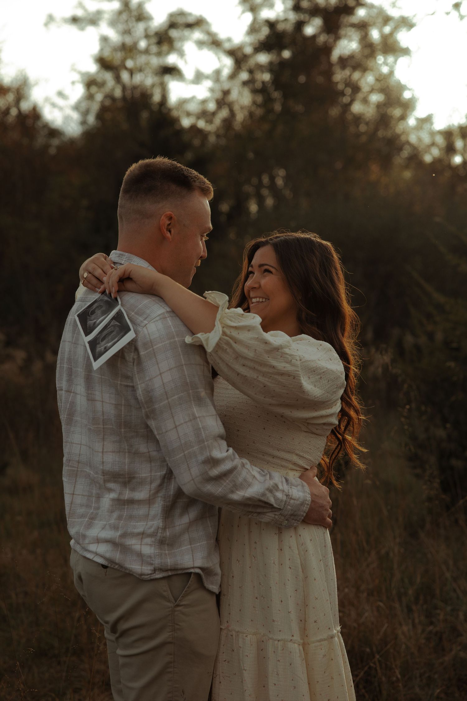Happy couple hugging in field holding ultrasound scan for romantic pregnancy reveal photoshoot.