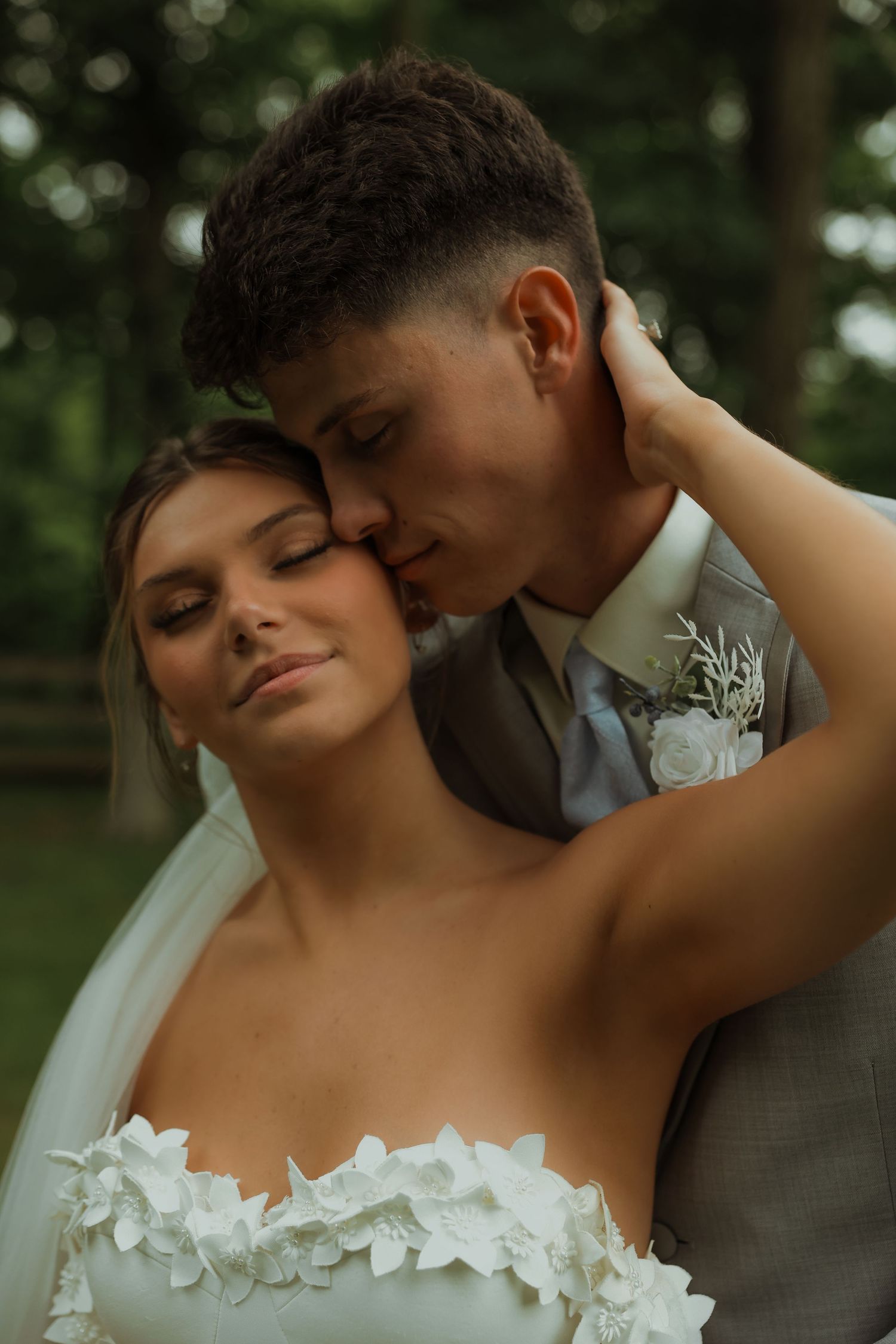 Bride with eyes closed smiles as groom kisses her cheek in romantic outdoor portrait.