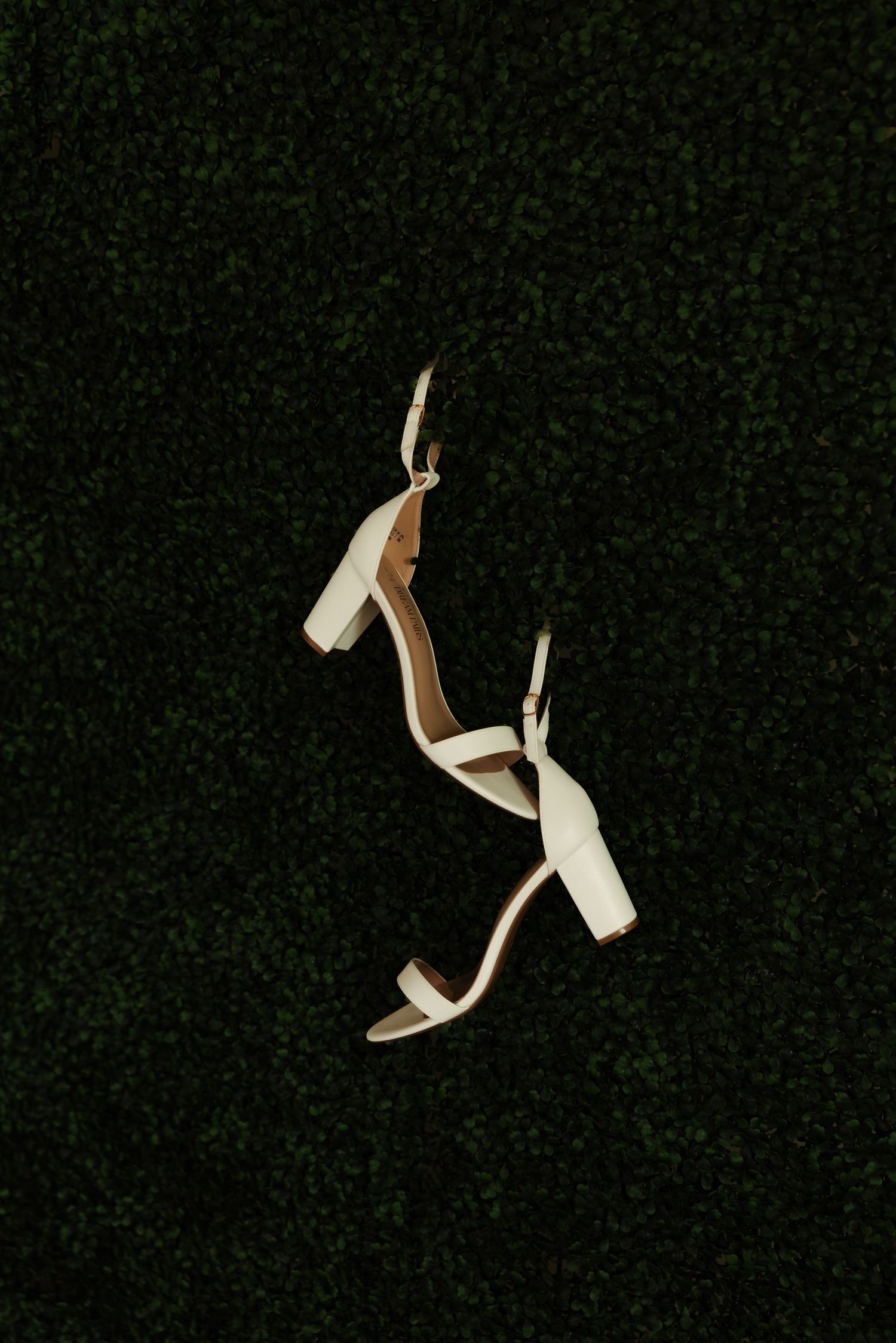 White block heel bridal sandals with ankle strap displayed against dark green foliage background.