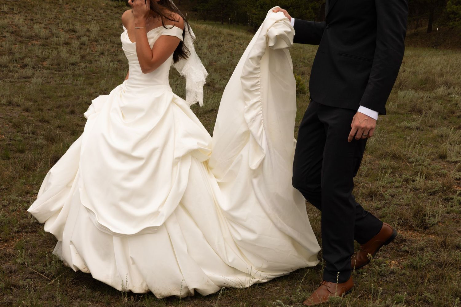 Bride in elegant off-shoulder ball gown wedding dress with groom in black suit holding her train outdoors.