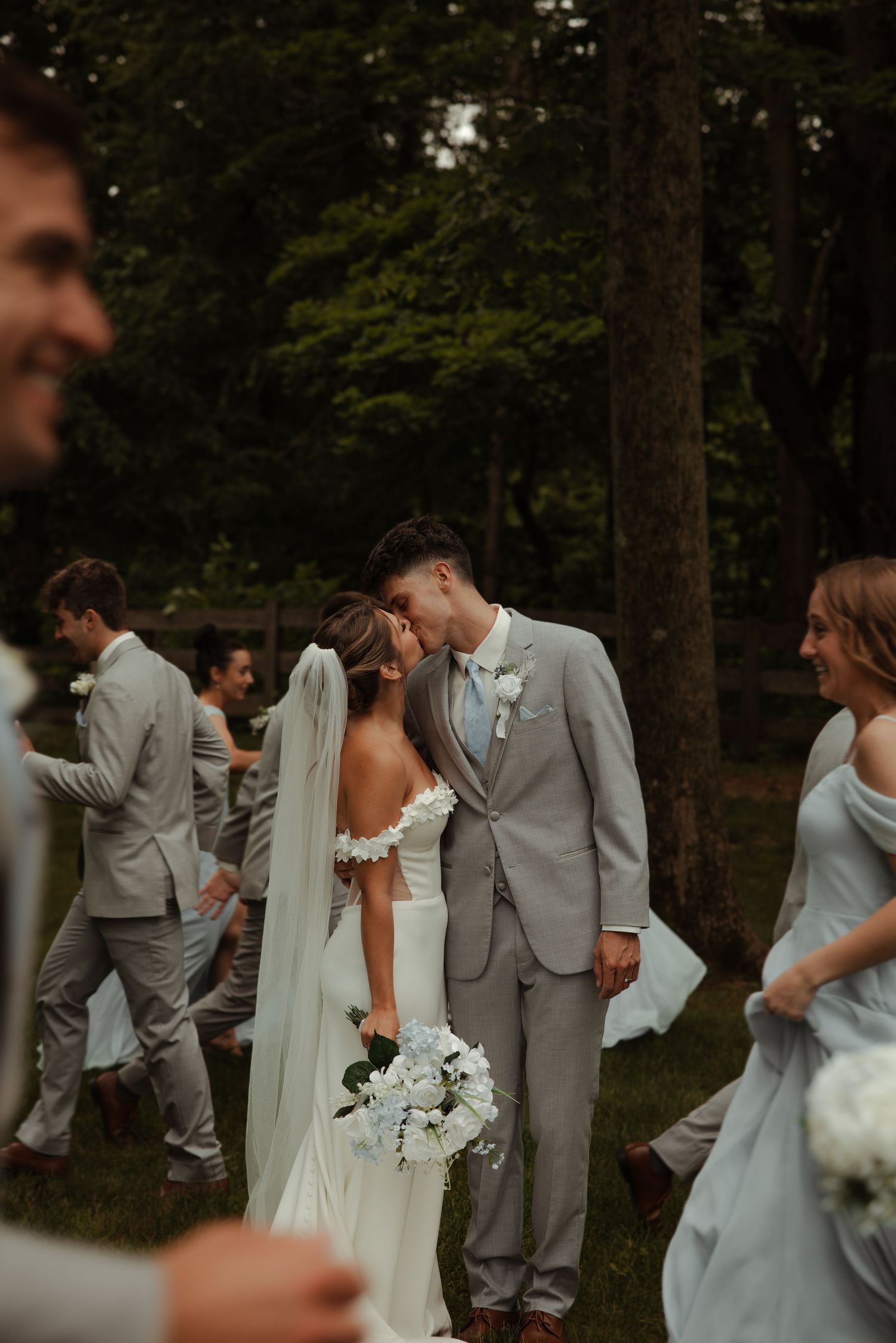 Bride and groom sharing a kiss surrounded by wedding party in gray and blue attire outdoors.