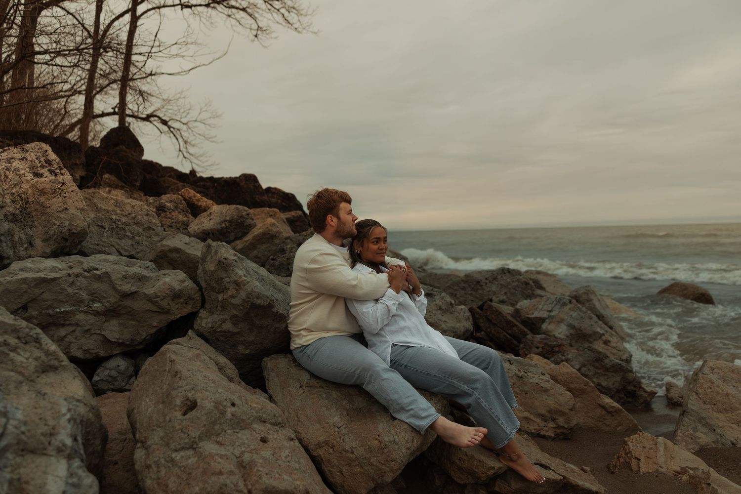 A couple sits together on rocky shoreline rocks gazing at the ocean under a cloudy sky.