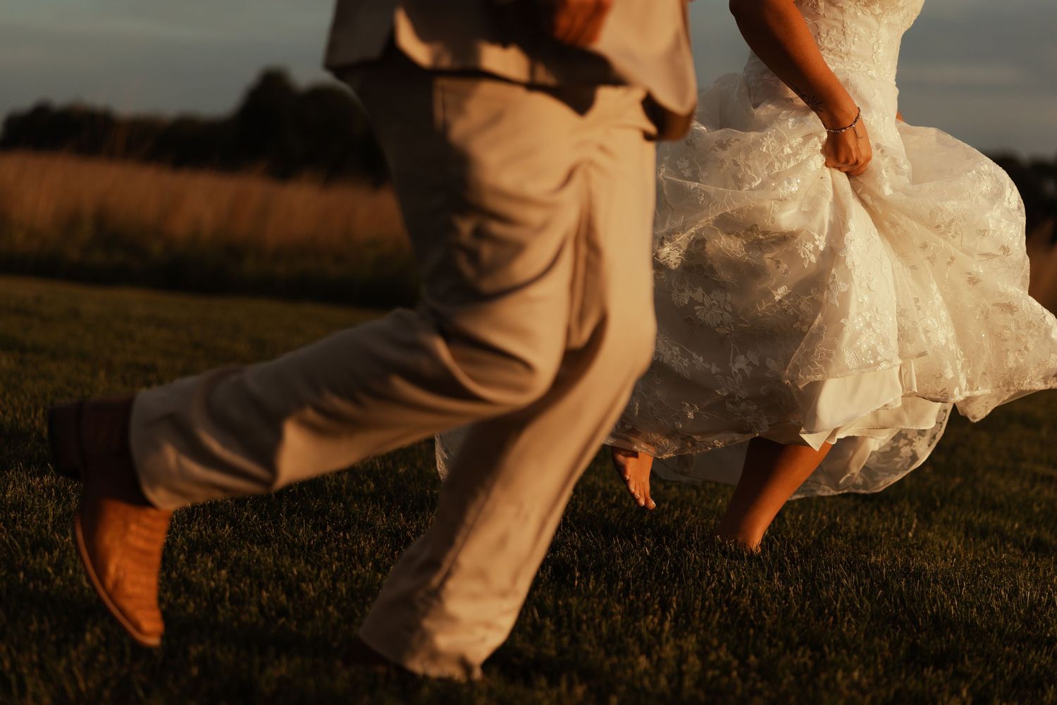 Bride and groom running through green grass field at golden hour, wedding dress flowing in motion.