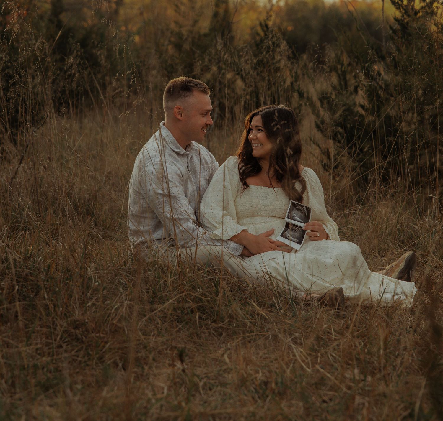 Couple sitting in a golden field holding an ultrasound photo during maternity session.