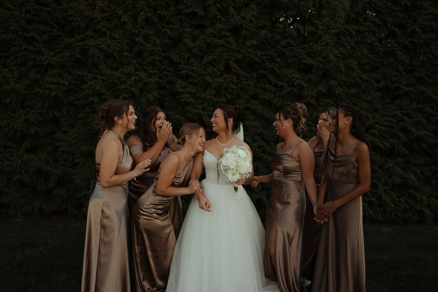 Bride in white gown laughs with six bridesmaids in gold satin dresses against a green hedge.