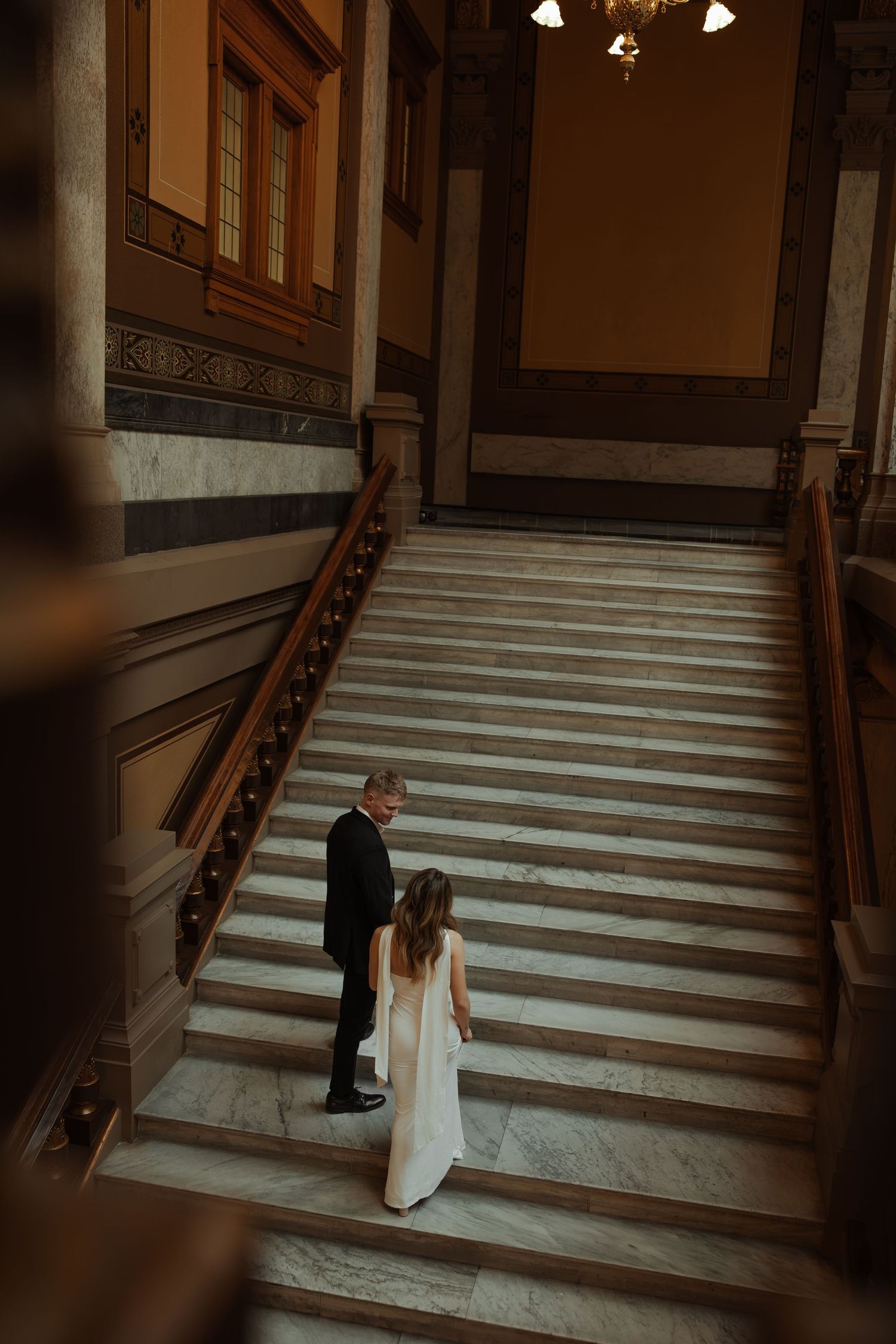 A bride and groom stand on marble stairs inside a grand historic building viewed from above.