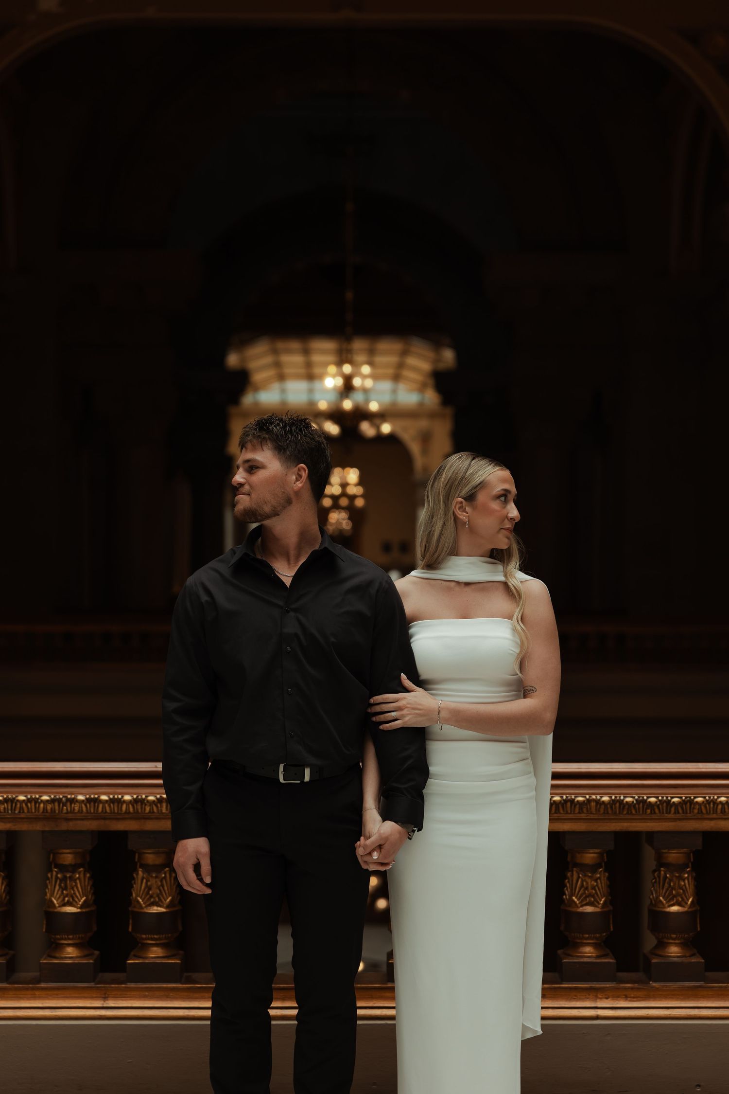 An elegant couple in formal attire holds hands inside a grand ornate building with chandeliers.