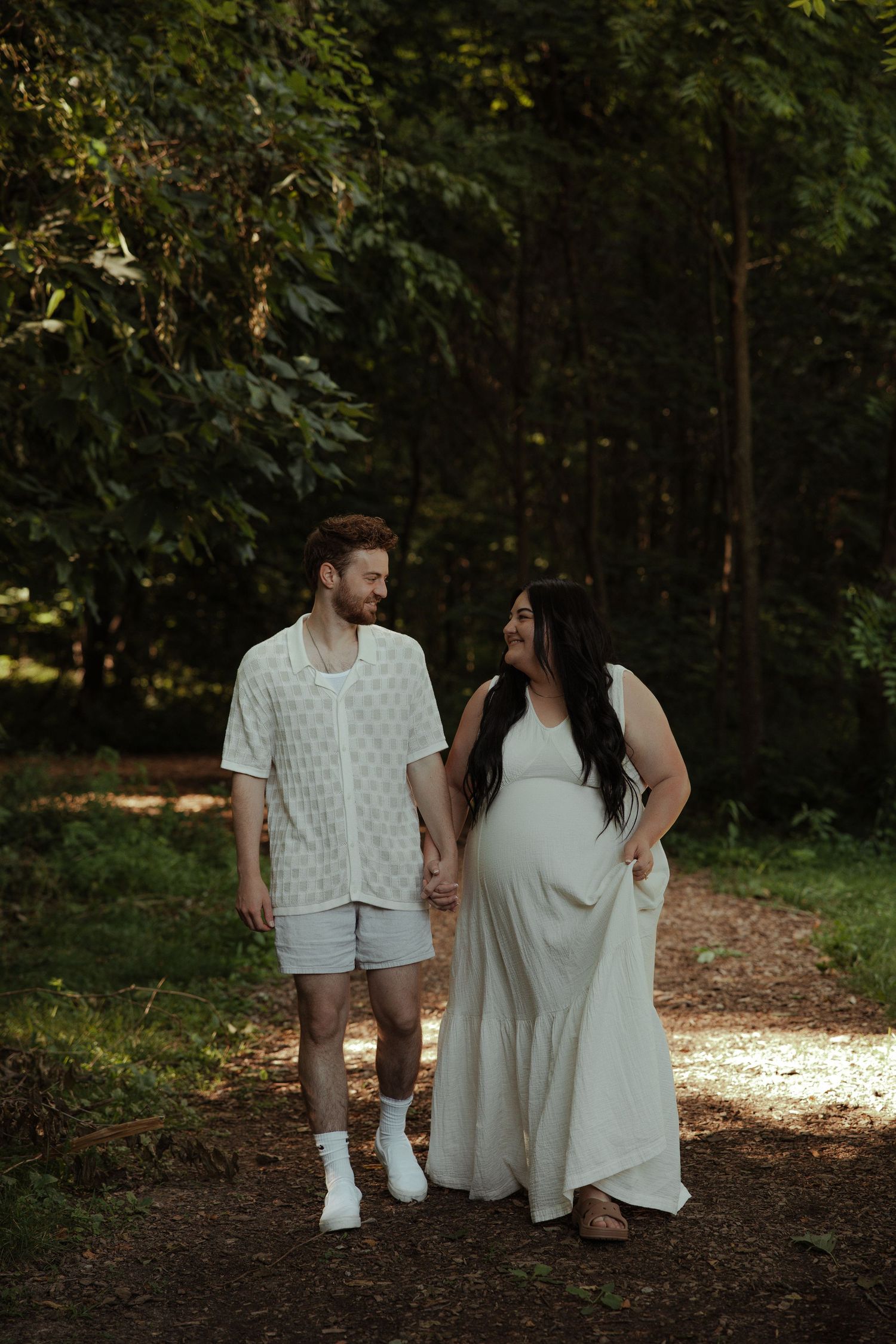 Smiling couple holding hands on a forest path, pregnant woman in flowing white dress with partner in casual wear.