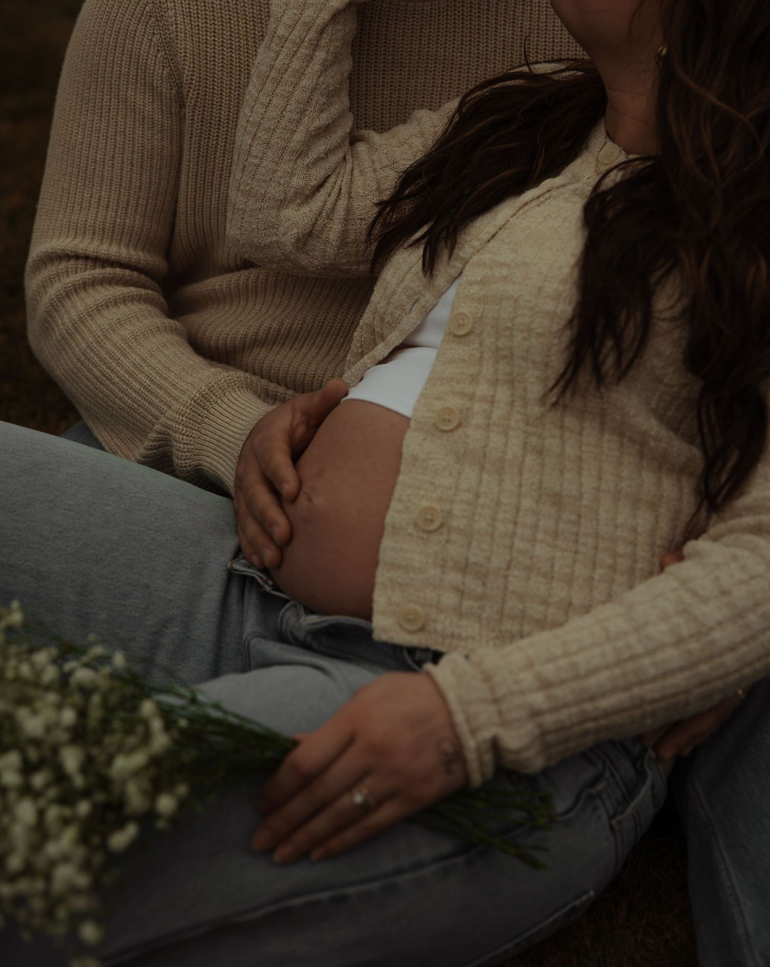 Pregnant woman in beige cardigan holds baby bump while partner embraces her outdoors with wildflowers nearby.