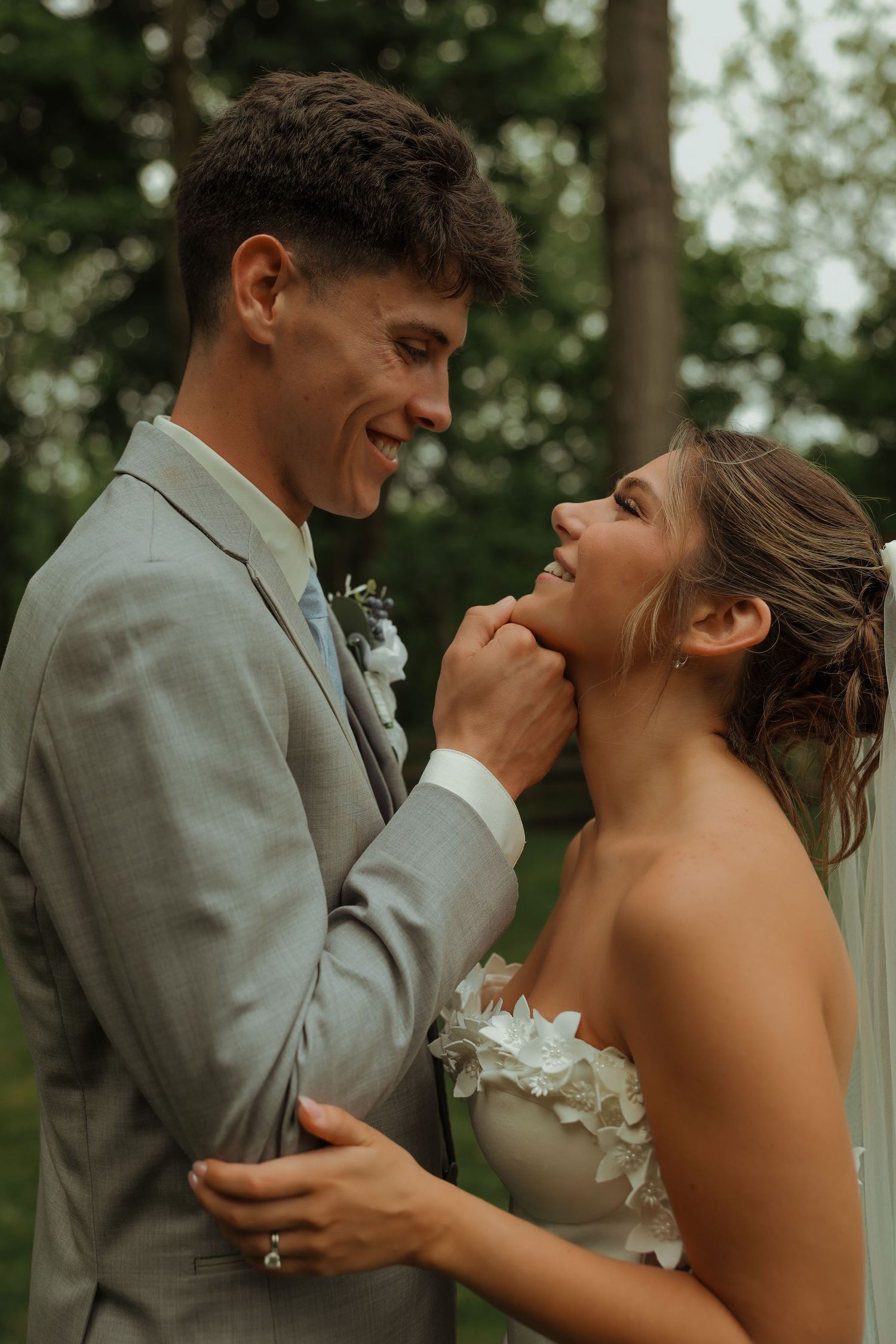 Bride and groom laugh joyfully together as he holds her chin in romantic garden portrait.