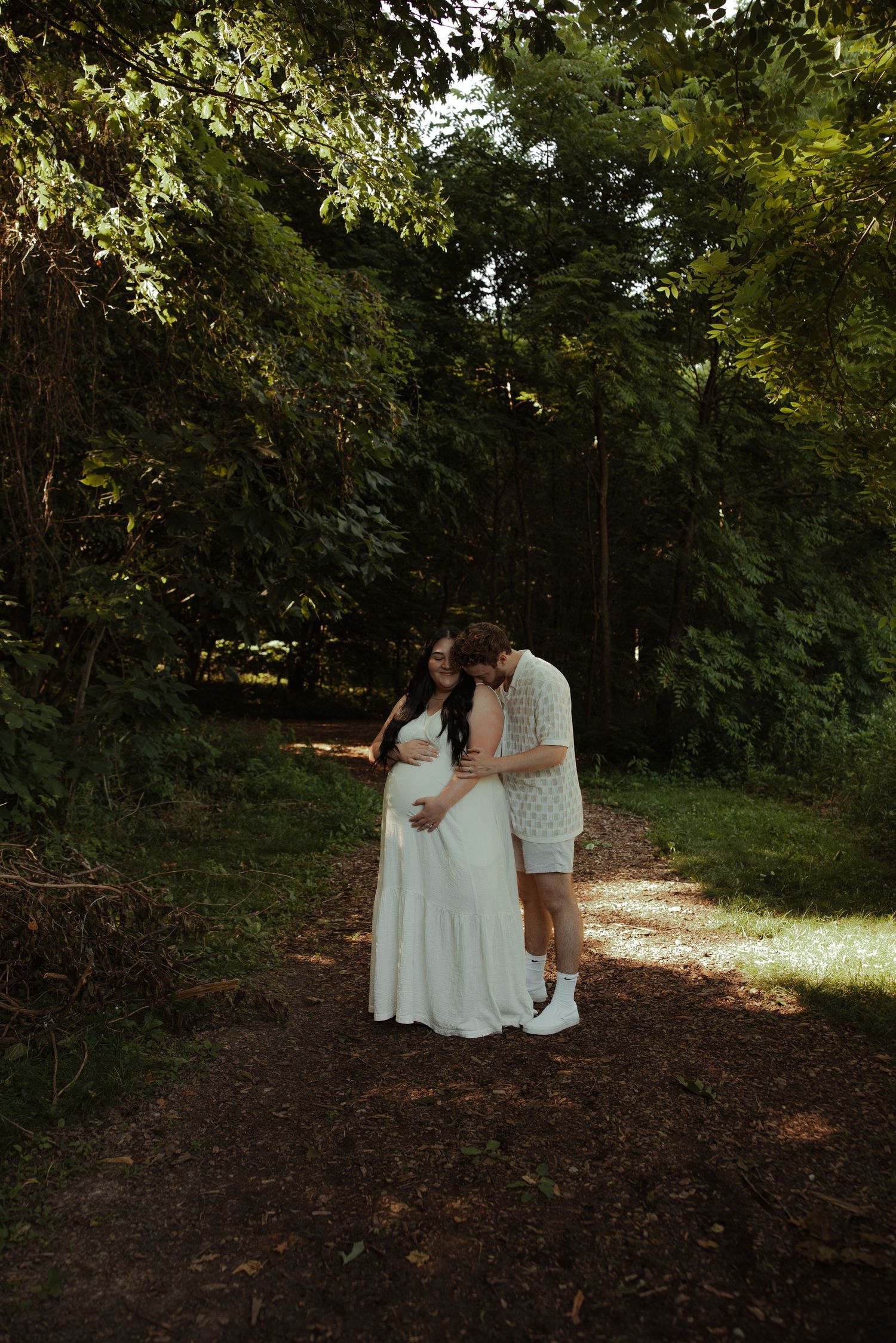 Couple on a forest path, man kissing pregnant woman's head as she cradles her bump in a white dress.