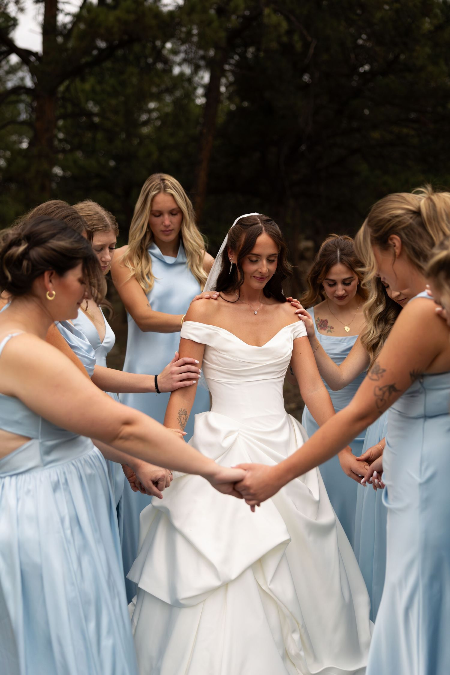 Bride in white gown surrounded by bridesmaids in light blue dresses holding hands in a circle outdoors.