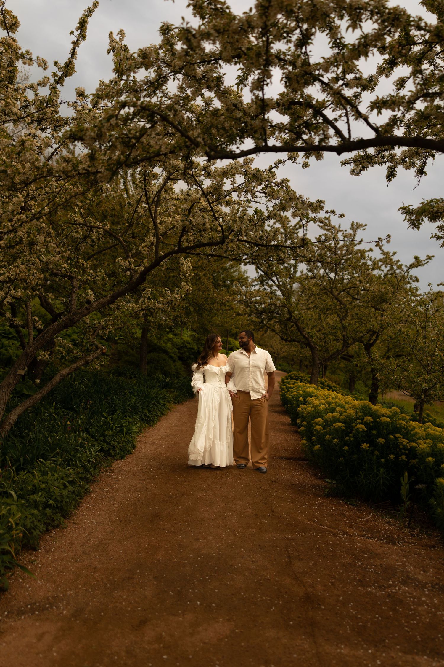 Couple in white wedding attire walking hand in hand along a dirt path lined with blooming trees.