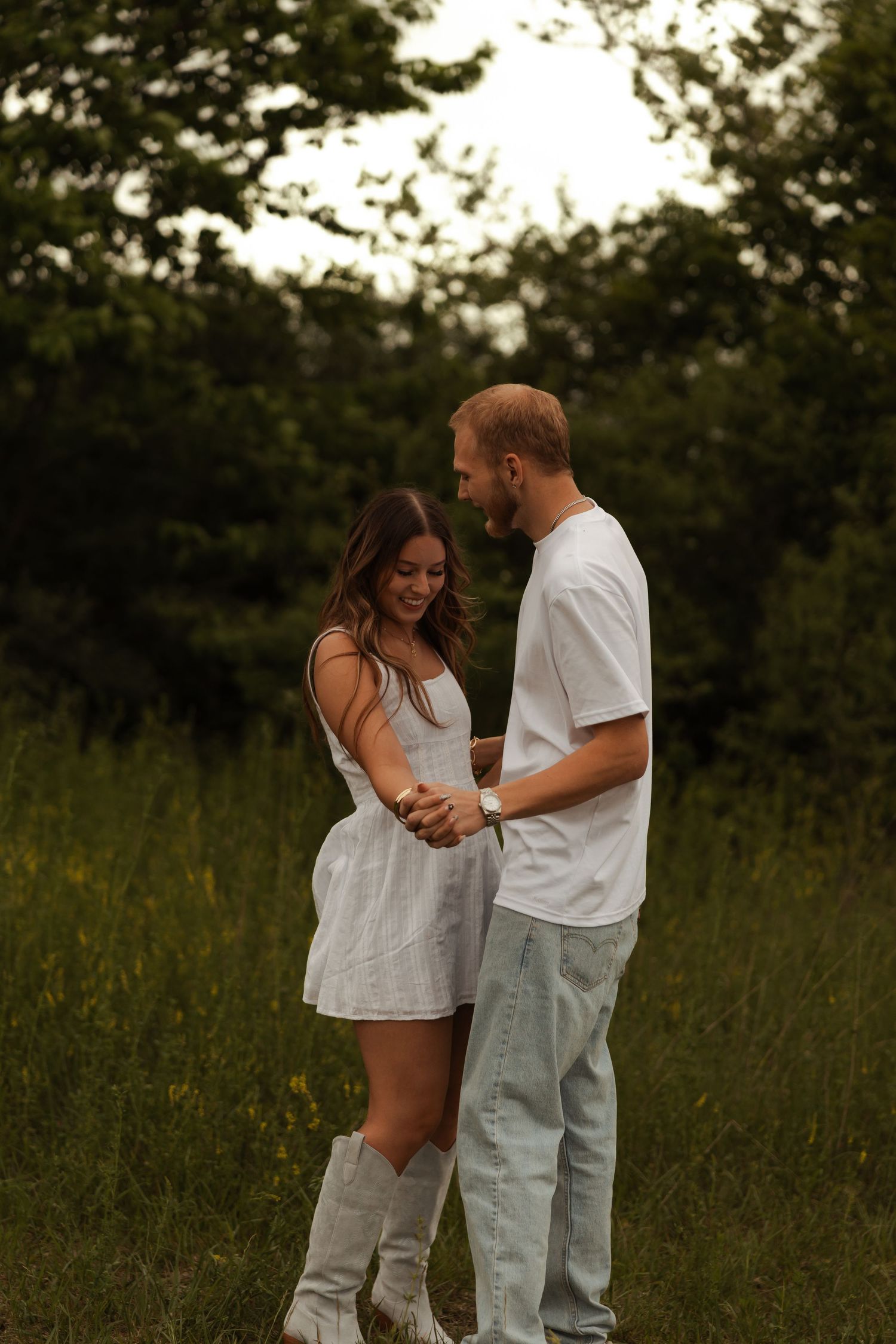 A smiling couple holds hands in a green field, the woman wearing a white dress and cowboy boots.