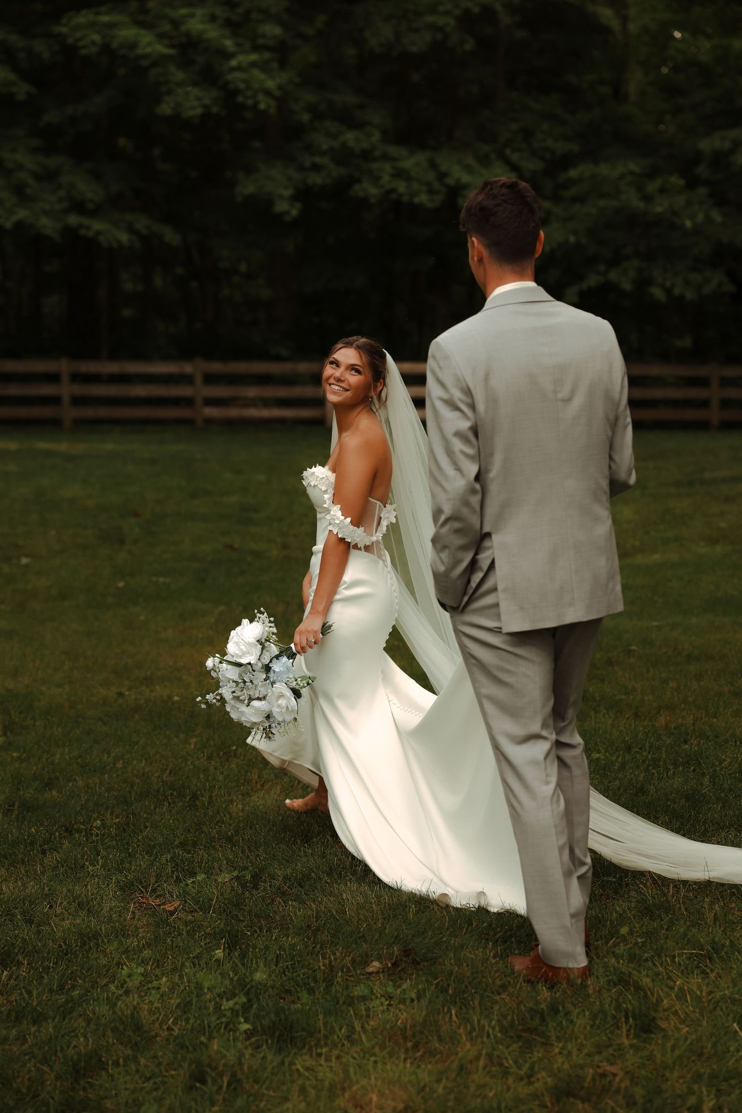 Smiling bride in a white gown holds a bouquet while walking hand-in-hand with groom on green lawn.