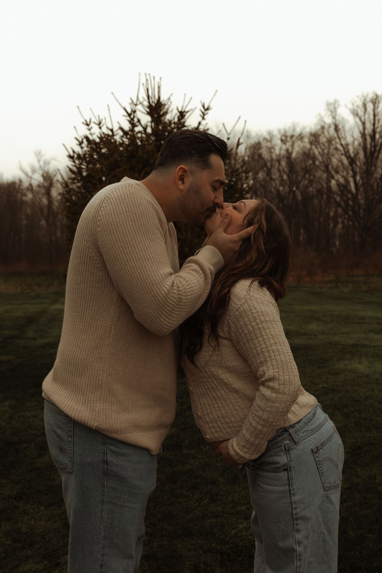 Couple sharing romantic kiss outdoors during cozy neutral-toned maternity photography session.
