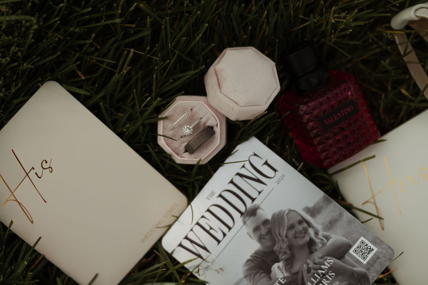 Wedding flat lay featuring ring box, perfume, wedding magazine, and vow book on grass.