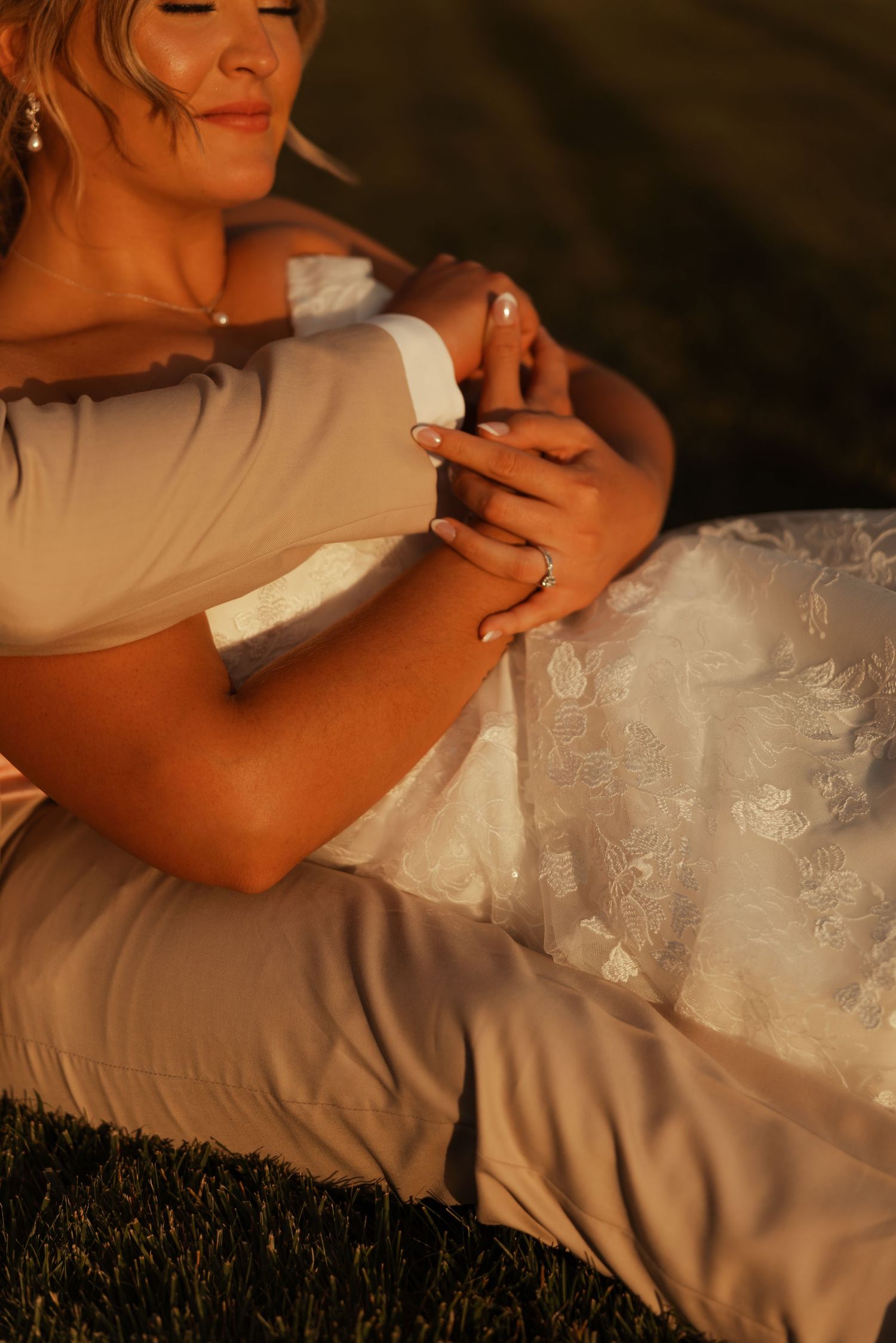 Groom embracing bride from behind, hands clasped, showing wedding ring in warm sunset light.