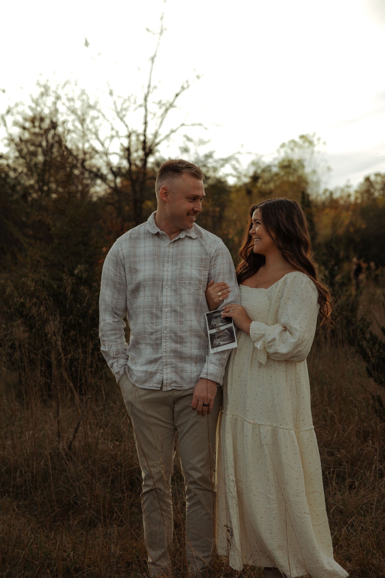 Smiling couple walking in autumn field holding ultrasound photos for pregnancy announcement session.