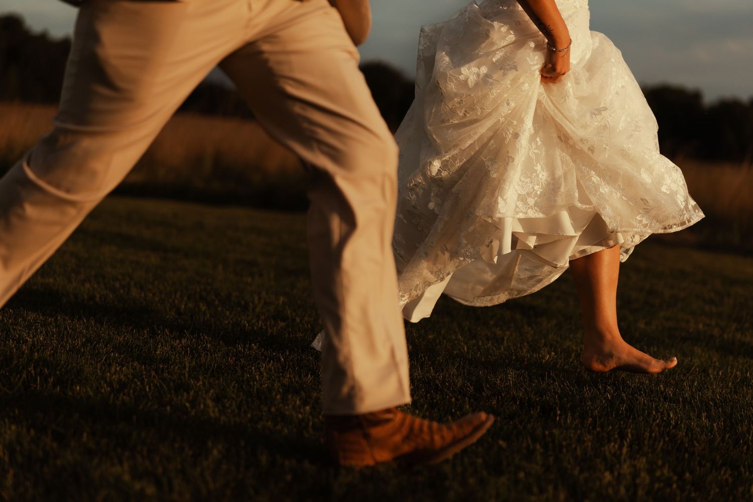 Bride and groom running barefoot through a sunlit field, wedding dress flowing in motion.