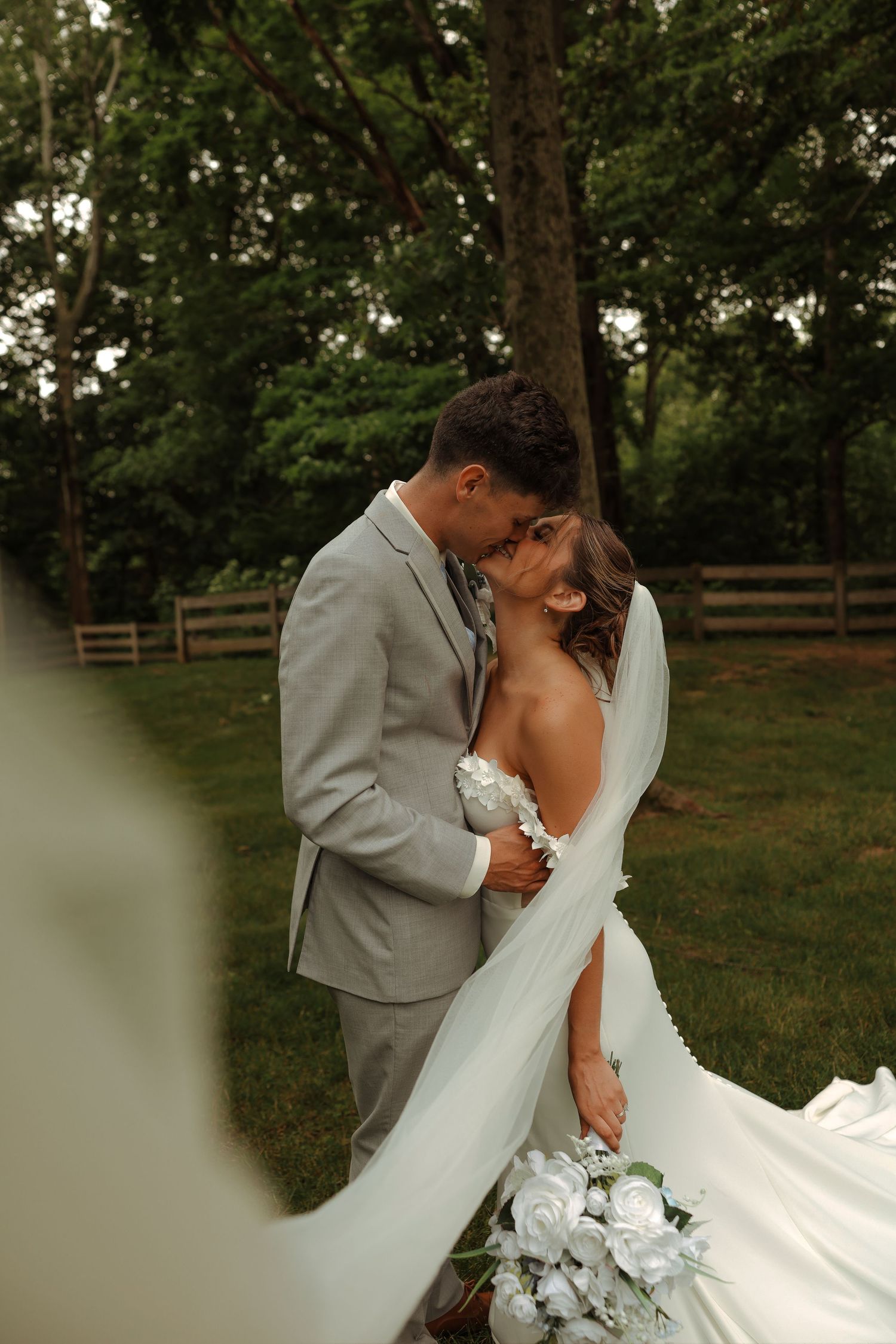 Bride and groom kiss romantically outdoors with white bouquet and flowing veil in garden.